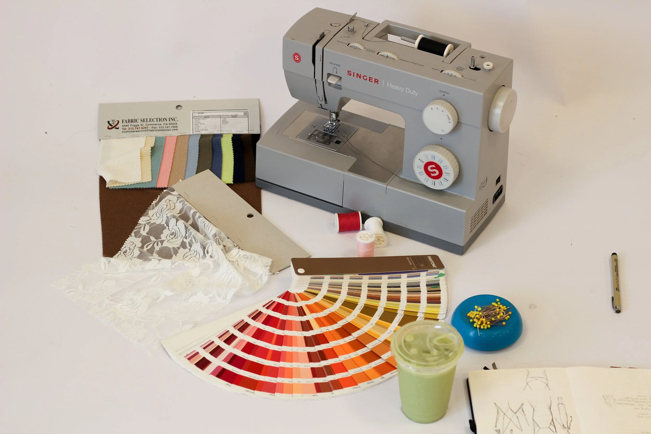 A collection of sewing and fabric design tools, including a sewing machine, fabric samples, color fan chart, buttons, sewing pattern book, and thread.