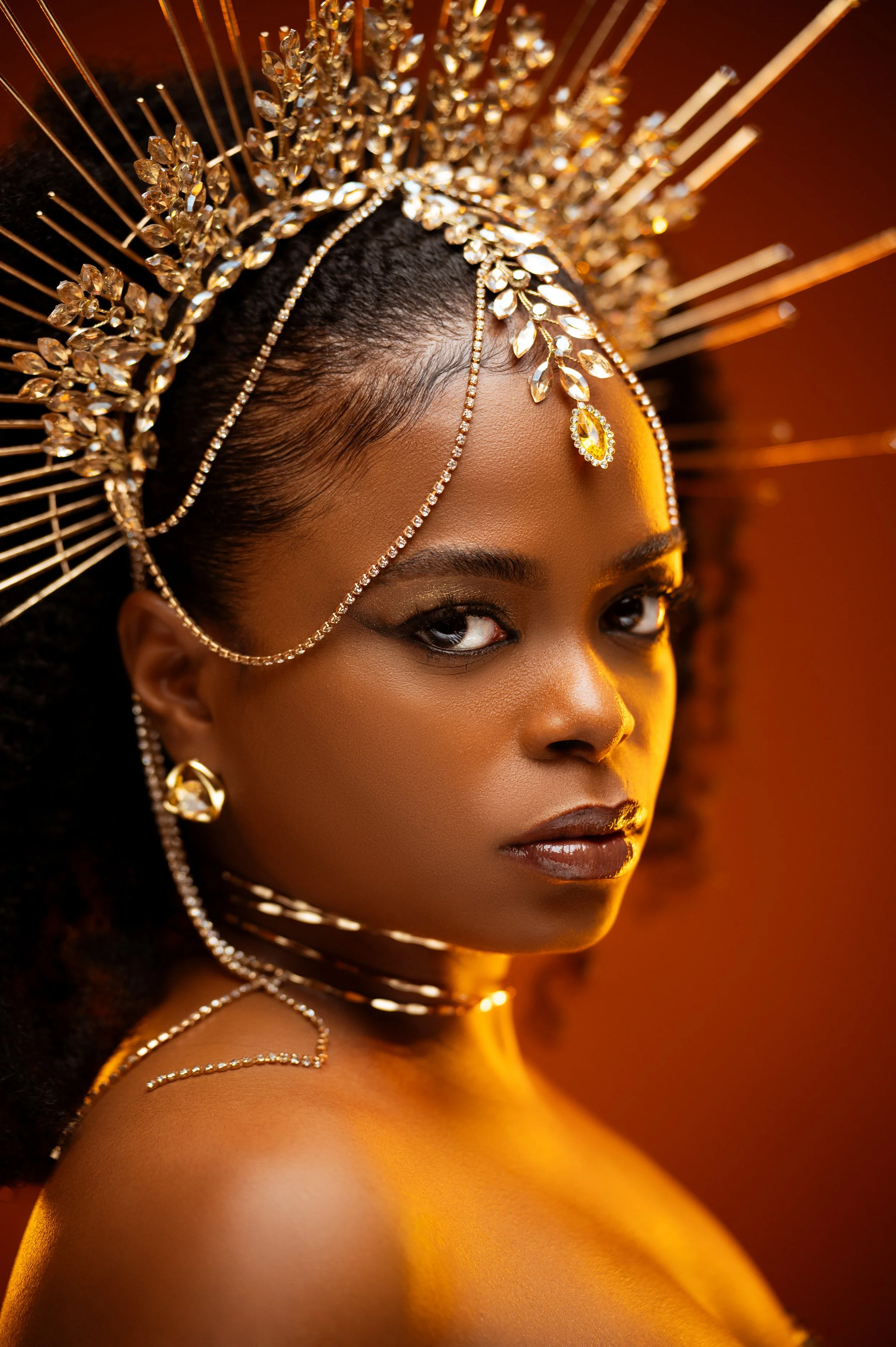Close-up of a woman with elaborate gold and crystal jewelry, including a large headdress with spikes and leaf-like decorations, and matching earrings and necklaces. The warm lighting highlights her skin and jewelry, with a focus on her eyes and lips.