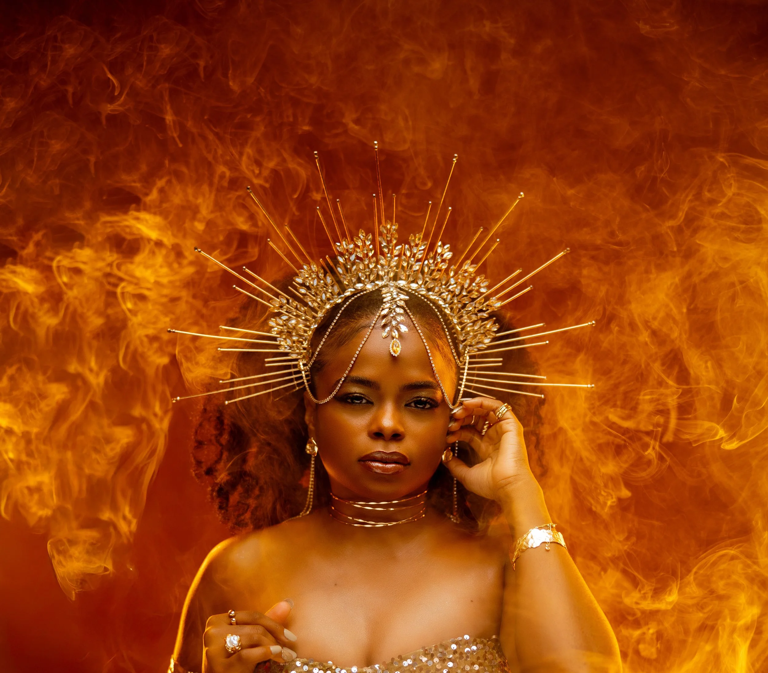 Portrait of a woman with a crown-like jewelry piece, in front of a fiery background.