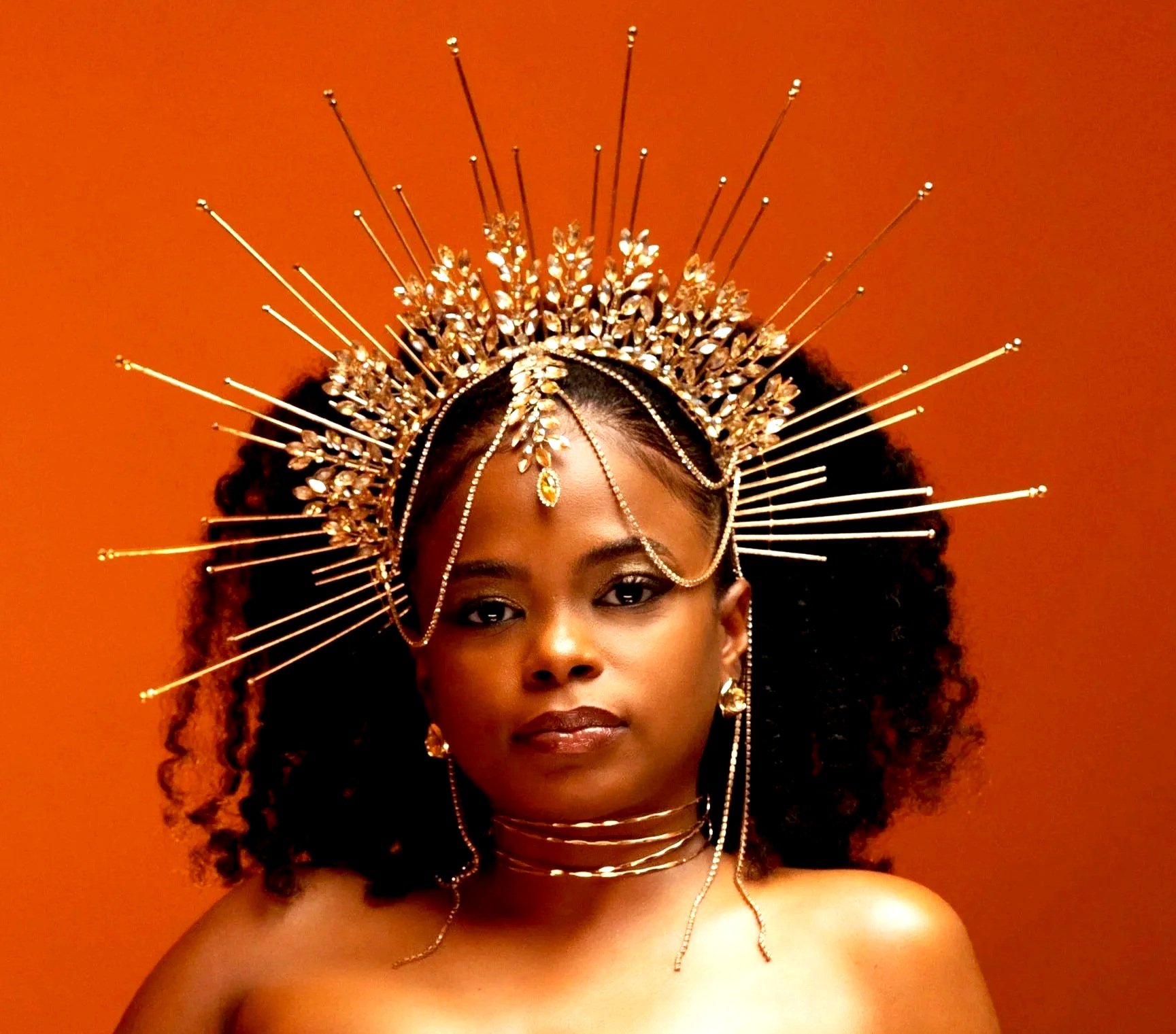 A woman with dark curly hair wearing an ornate gold and crystal crown and jewelry, posed against an orange background.
