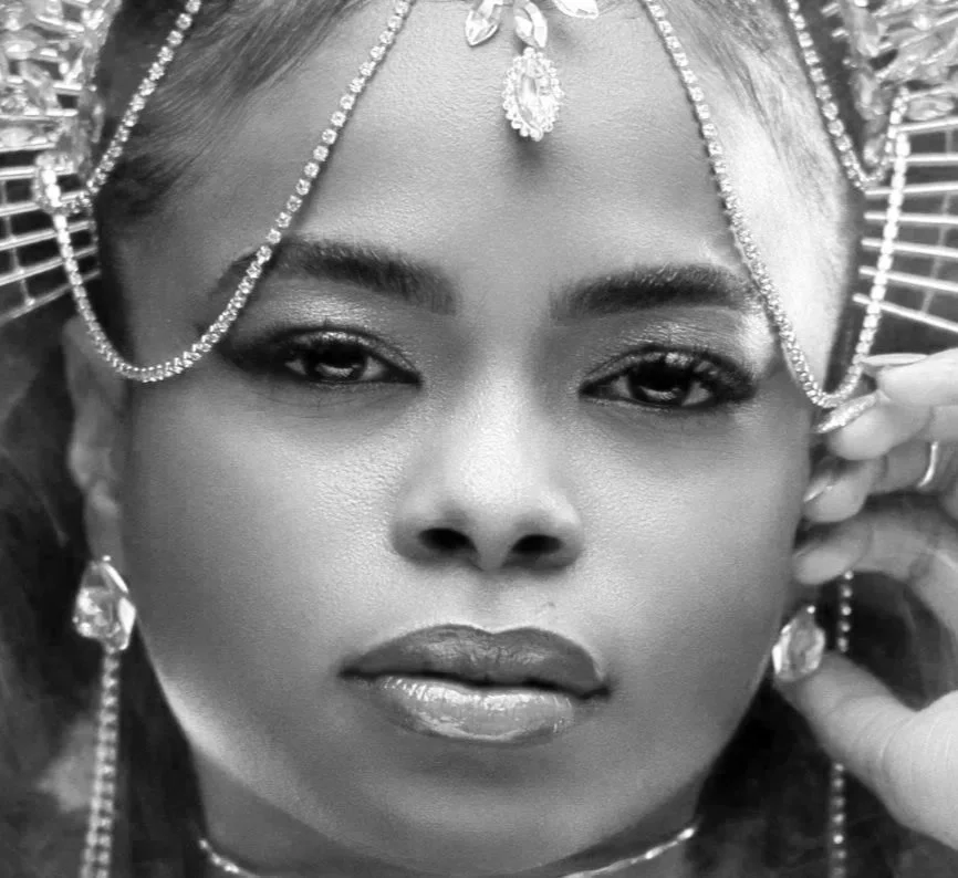 Close-up black and white photo of a woman wearing elaborate jewelry and headpiece, with detailed eye makeup and glossy lips.
