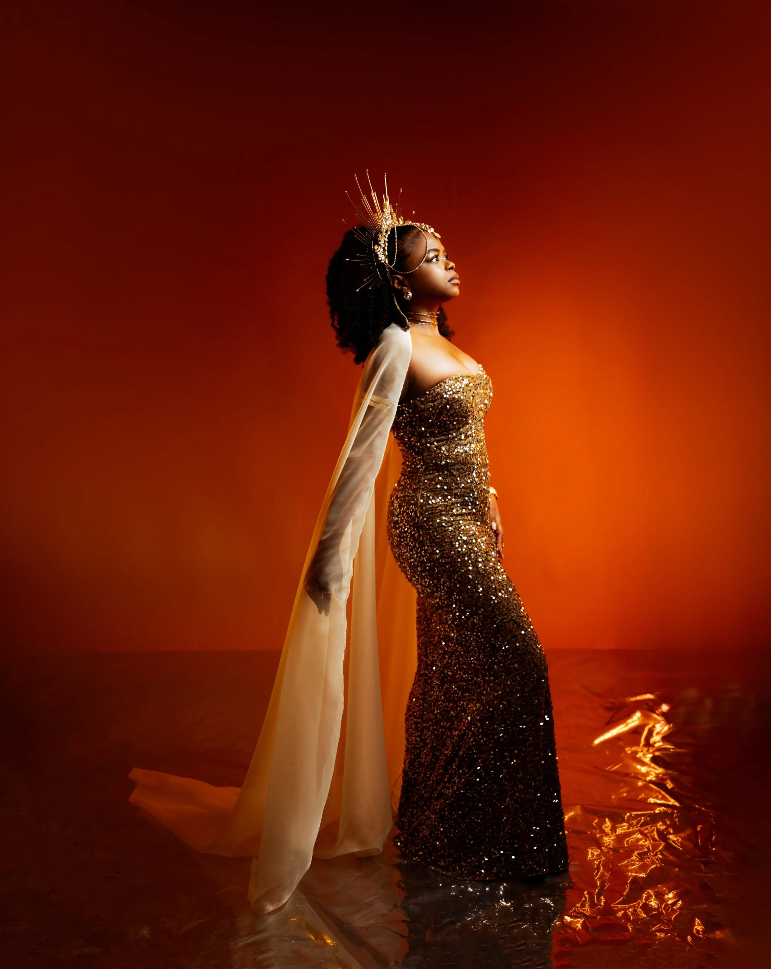 A woman in a gold sequined dress with a sheer cape and an ornate gold headpiece stands against an orange background with reflections on the floor.