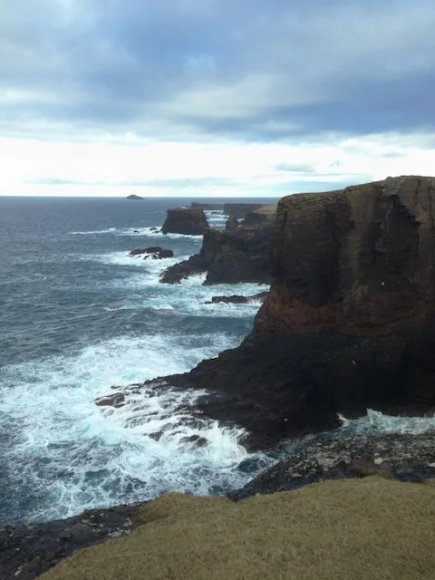 A portrait aerial photo of the dramatic east coast of Scotland. Waves crashing against the rugged shoreline
