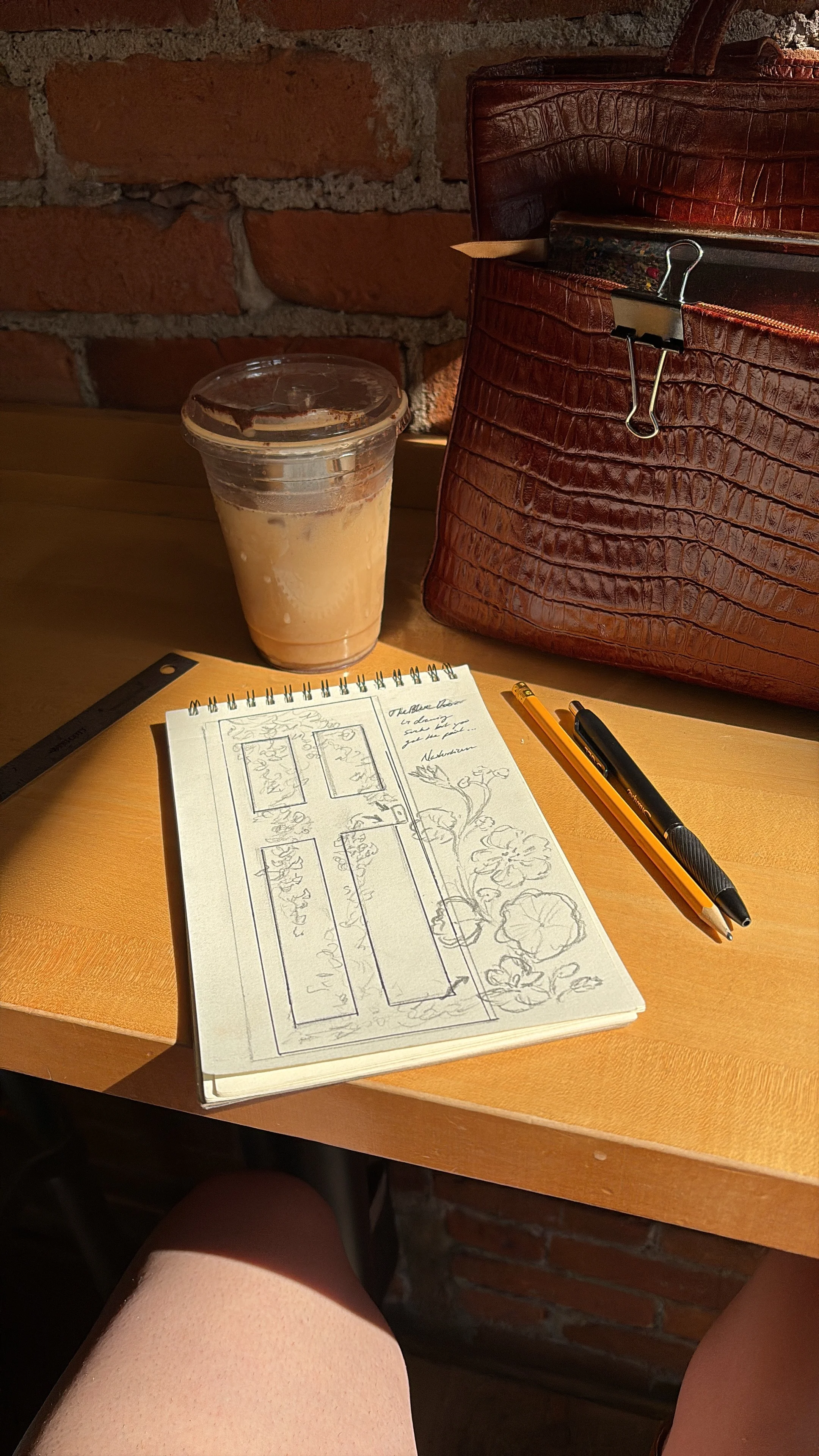 A sketchbook with a garden layout drawing, a black and orange pen, and a yellow mechanical pencil on a wooden table. A large brown leather bag and a plastic cup with a cold beverage are on the table, with a brick wall in the background.