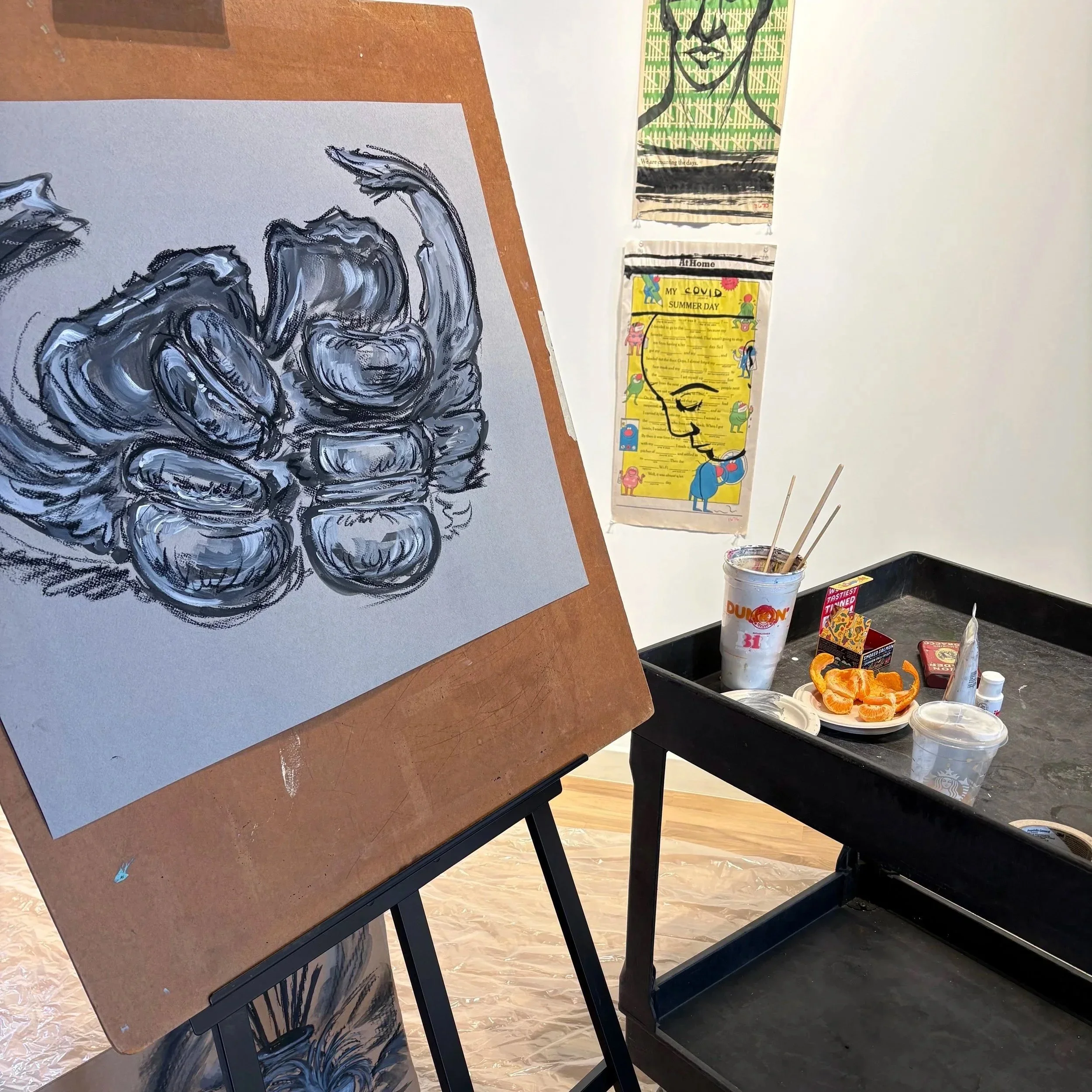Part of an art studio with a wooden table, an abstract black and white painting on paper, various art supplies including a Dunkin' drink cup with paintbrushes, and colorful wall hangings, including one with cartoon characters and one with a stylized face.