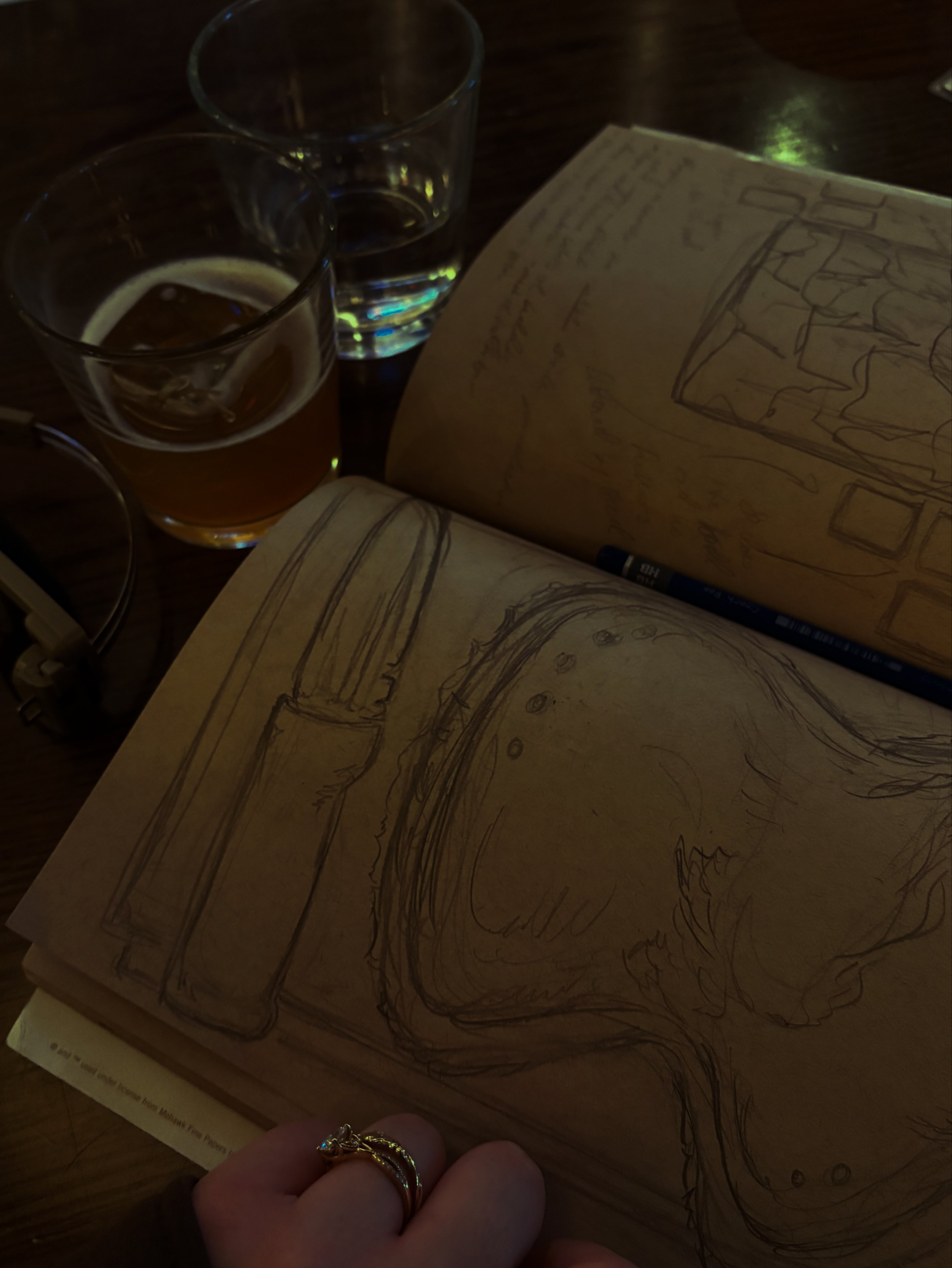 Close-up of a person's hand holding a gold ring, with sketchbooks and drawings of a lamp and a cabinet on the table, and two glasses, one filled with water and the other with beer, in a dimly lit setting.