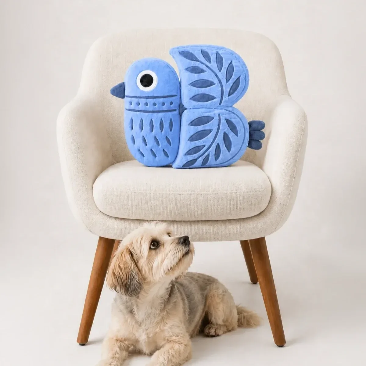 A white armchair with wooden legs features a large, plush blue butterfly-shaped pillow on it. A small, light-colored dog with floppy ears is sitting underneath, looking up at the pillow.
