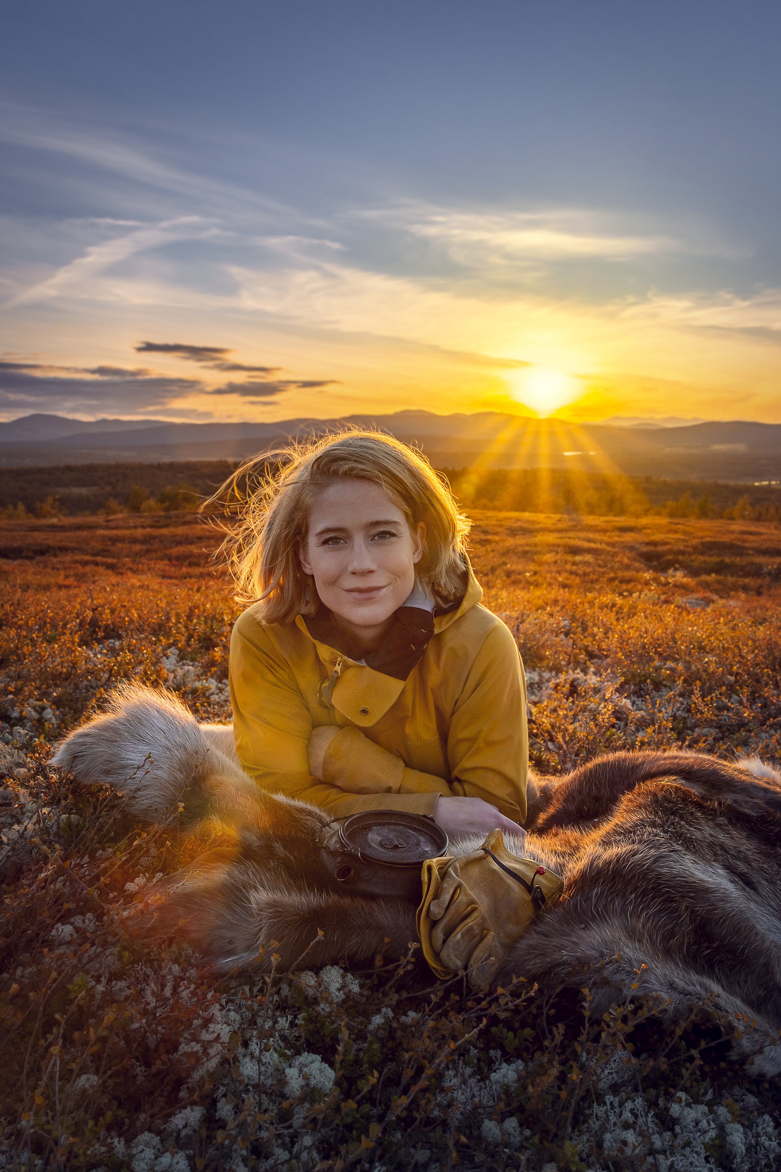 En kvinne i en gul jakke som ligger på en frossen jord med gress og lav vegetation, solnedgang i bakgrunnen, omgitt av hunder, på en ås eller fjellområde.