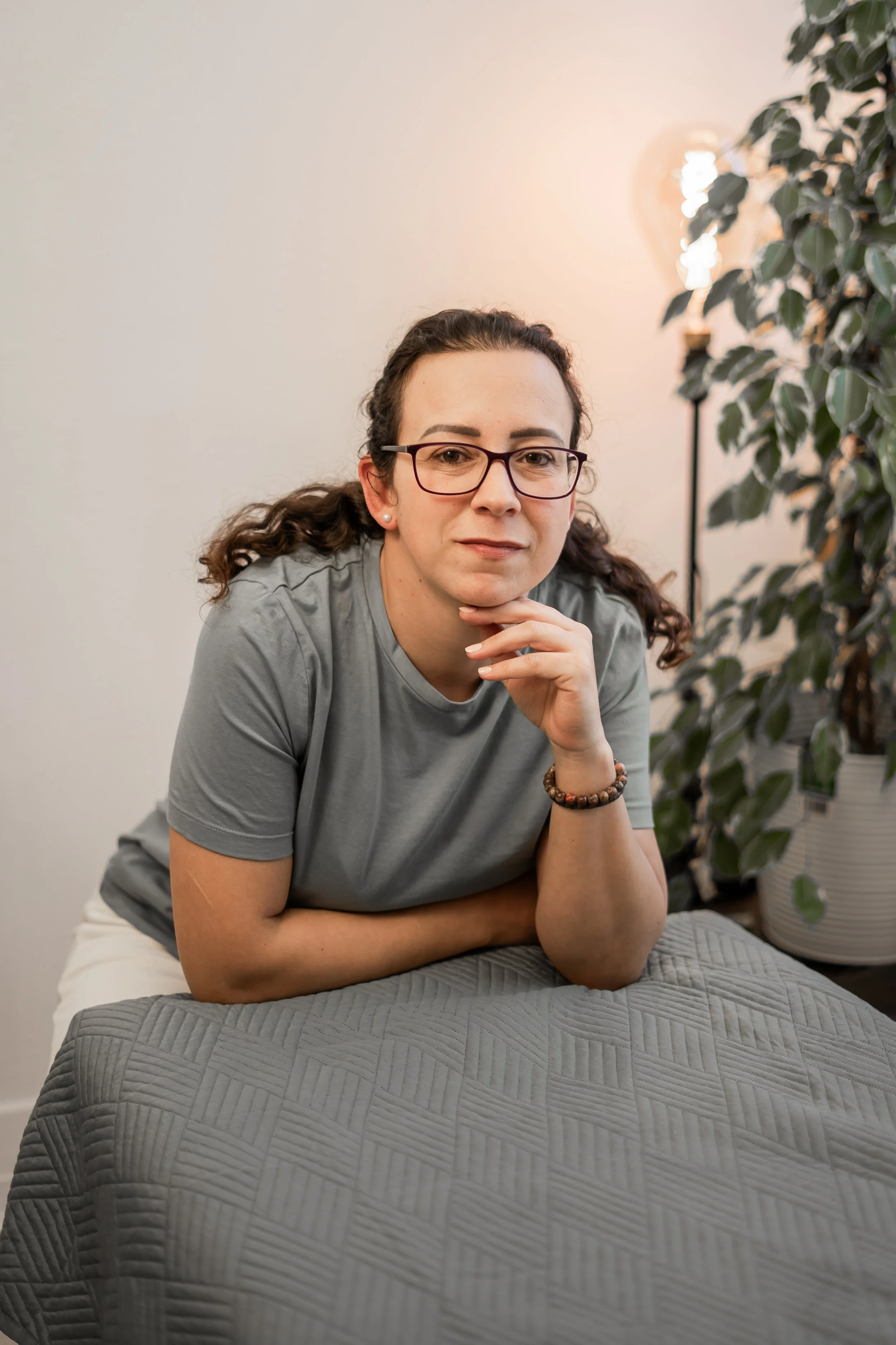 A woman with glasses and dark curly hair, wearing a gray t-shirt, sitting on a bed with a quilted gray bedspread, resting her chin on her hand, with a large potted plant and a lamp in the background.