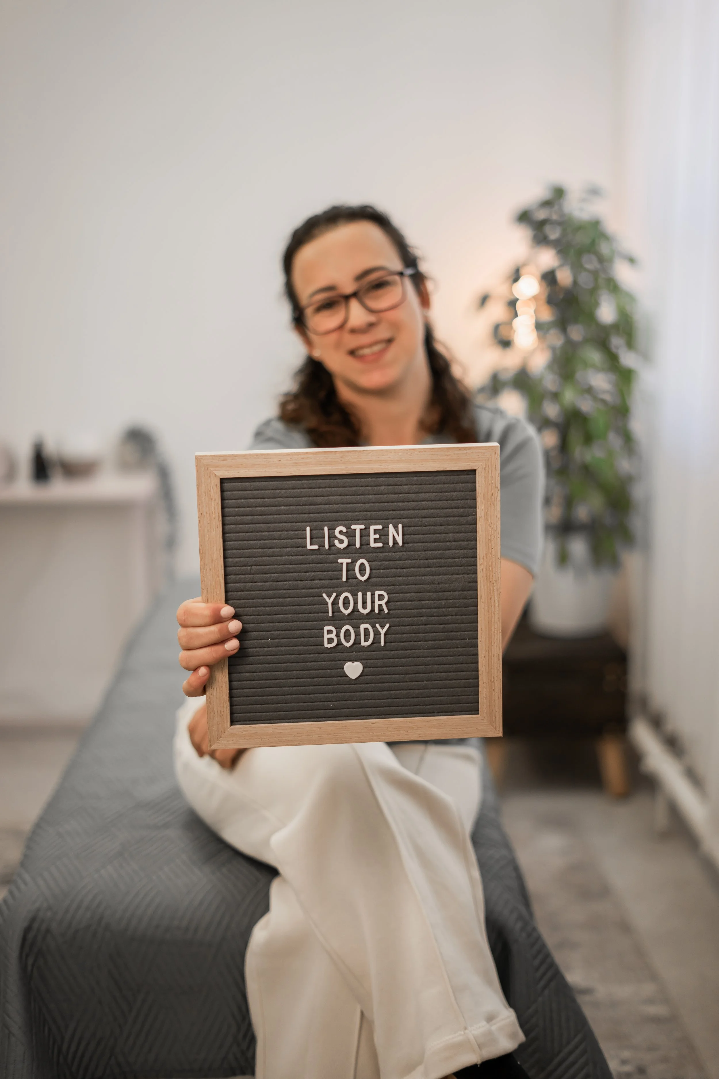 A woman with glasses, dark hair, and a gray shirt, sitting on a gray chair, holds a message board that reads 'LISTEN TO YOUR BODY' with a small heart symbol. There is a green plant and a window in the background.