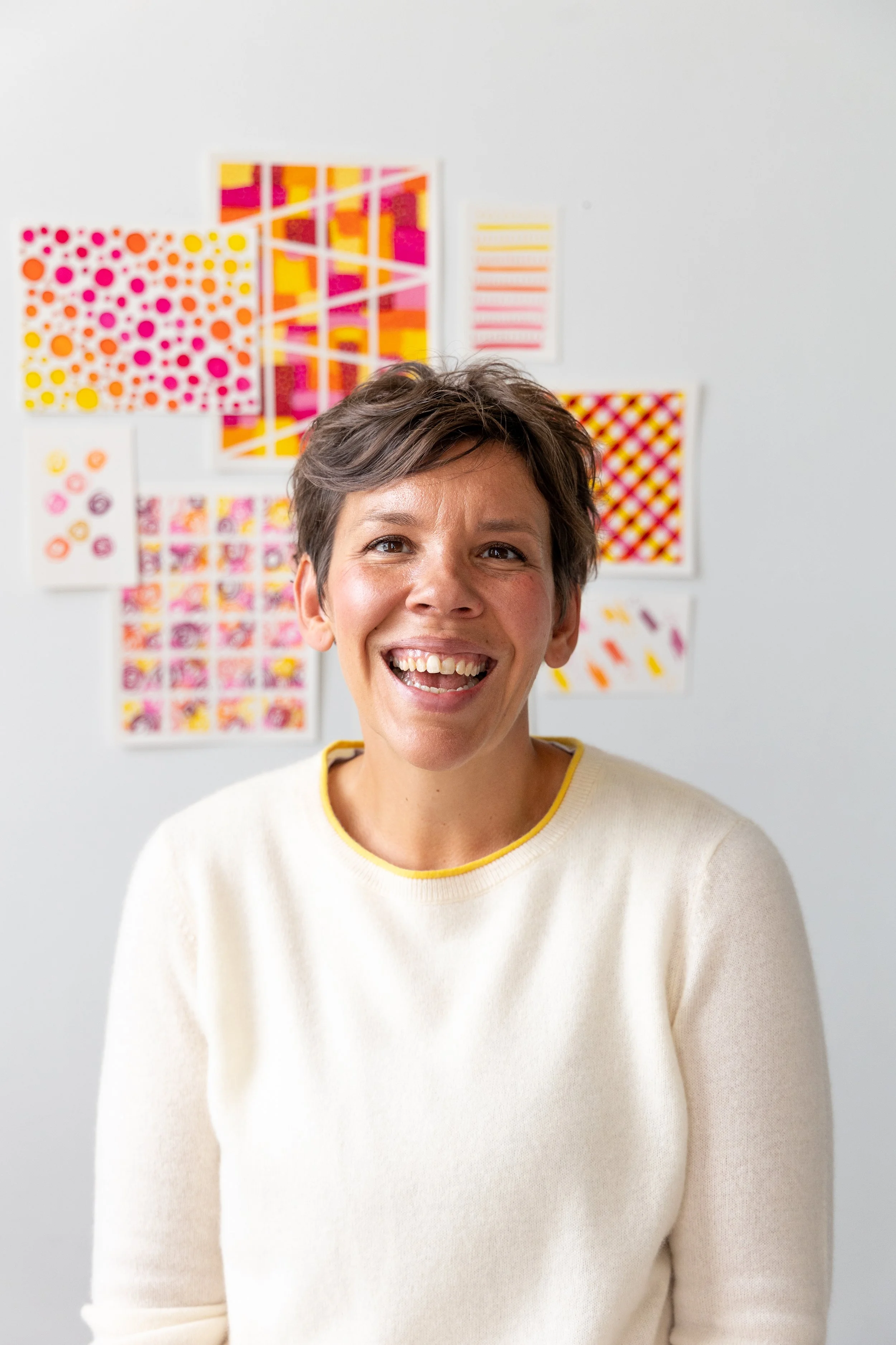 Picture of Emily sitting in front of a wall where her art is hanging. She is wearing an ivory sweater with yellow trim and has a big smile on her face.