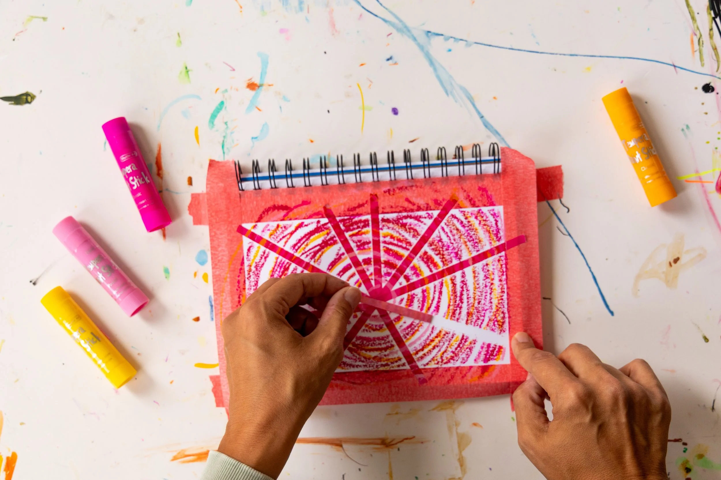 A picture of Emily peeling off washi tape from a page in her art journal. There are tempera paint sticks near the notebook.