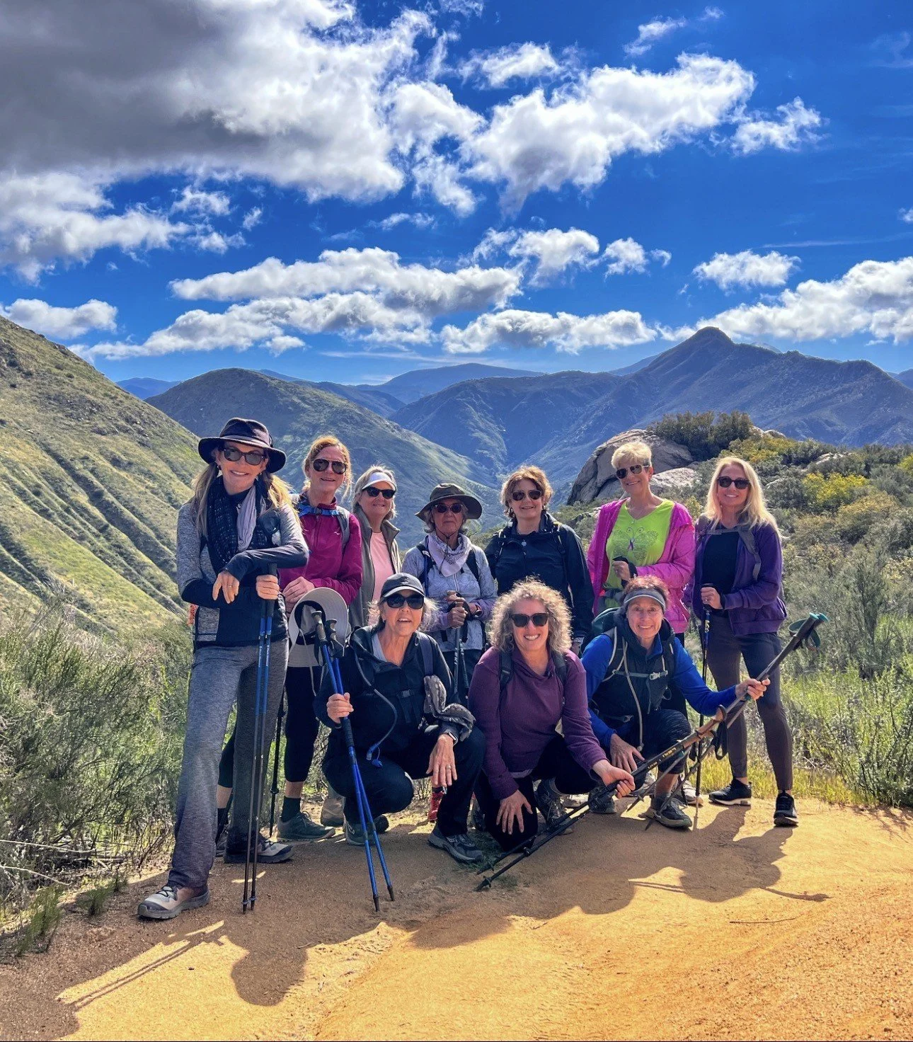 A group of ten women hiking on a trail in a mountainous landscape under a partly cloudy sky.