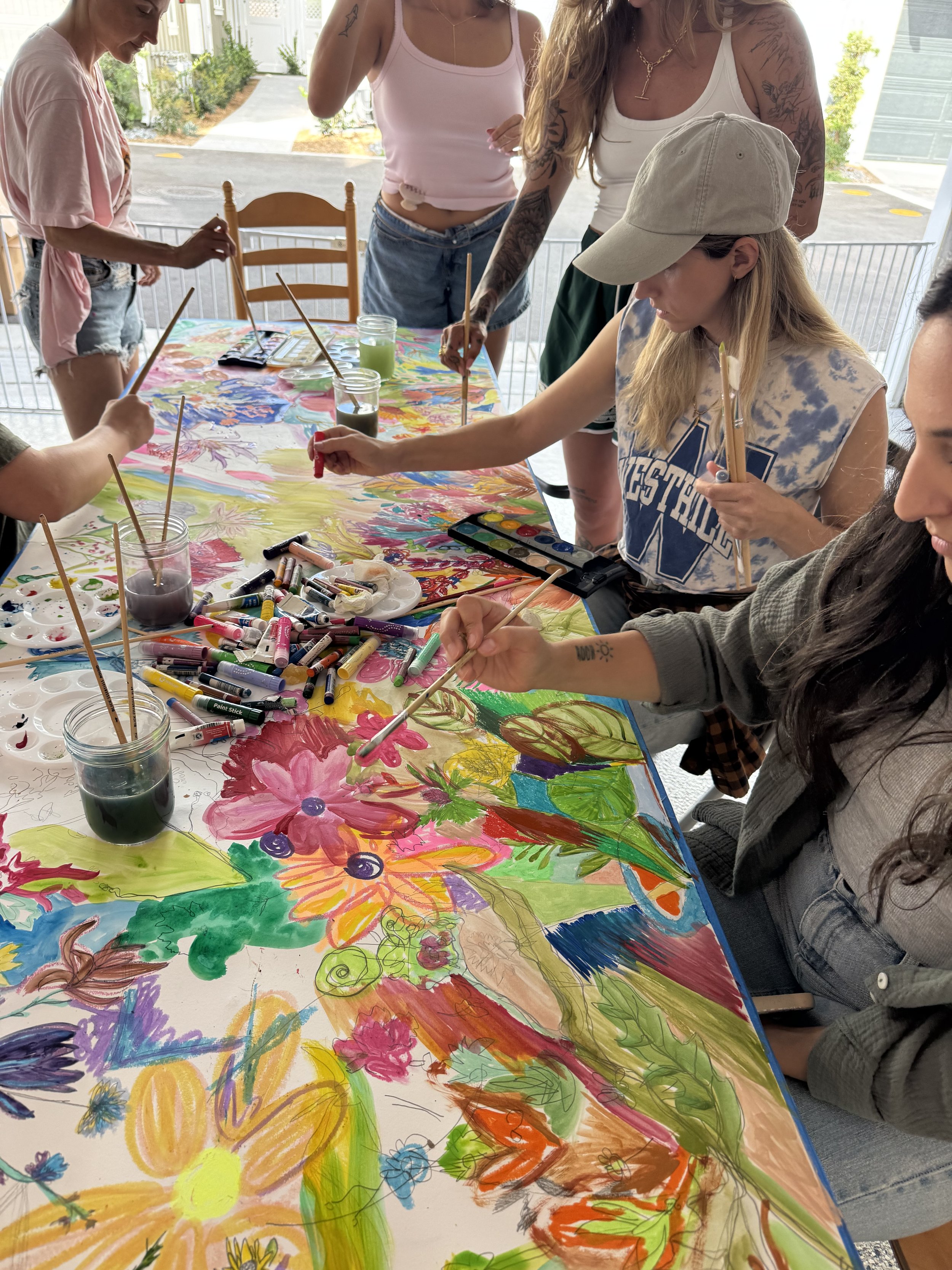 People are gathered around a table painting a large colorful mural with flowers and abstract designs using markers and watercolors inside a bright room.