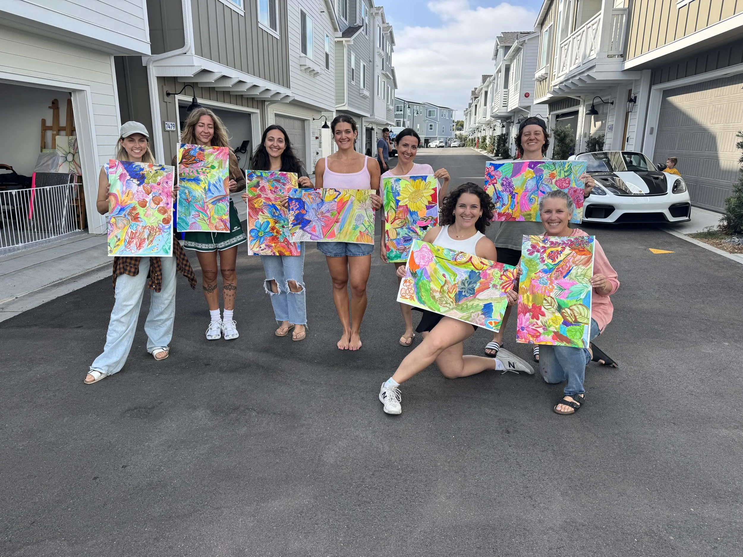 A group of nine women holding colorful abstract paintings standing in a parking lot in front of multi-story residential buildings, with two cars and a young boy visible in the background.