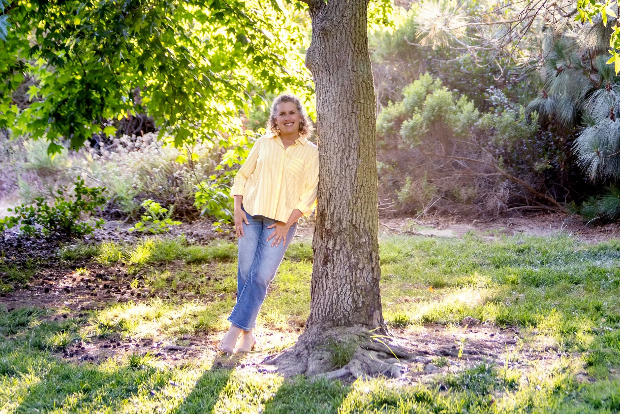 A woman with curly hair wearing a yellow striped shirt and jeans, standing behind a tree in a lush green outdoor area, smiling and looking at the camera, surrounded by trees and sunlight.