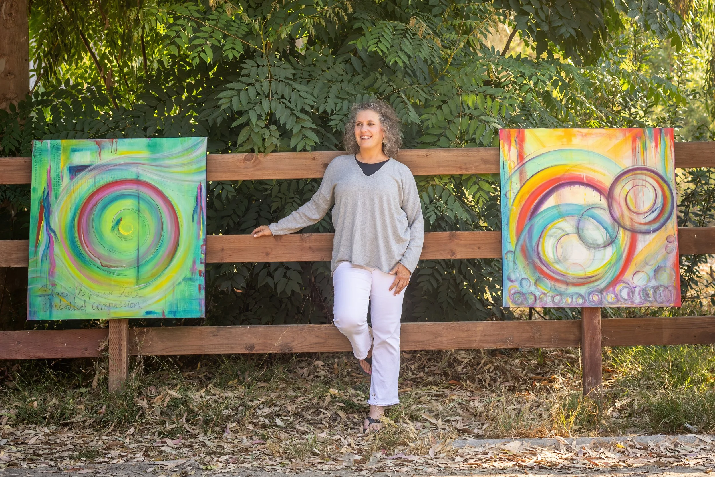 Denise Joy standing outdoors next to a wooden fence with colorful, abstract spiral paintings.