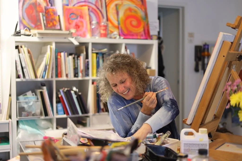 A woman enjoying her painting in an art studio, surrounded by colorful artworks, books, and art supplies.