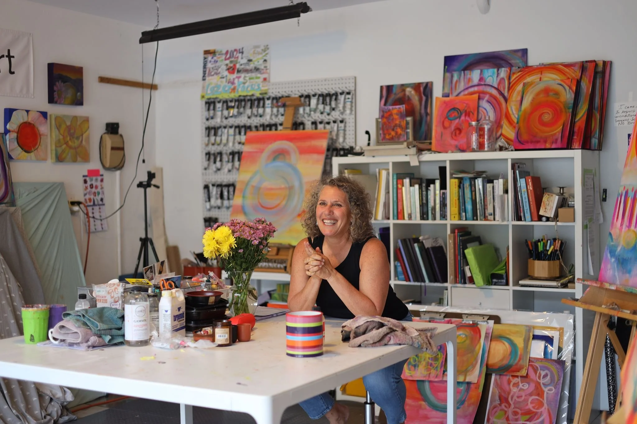 A woman with curly hair and a black sleeveless top smiling and sitting at an art studio table with a bouquet of flowers in front of her, surrounded by art supplies and completed abstract paintings with bright colors and swirling patterns on the walls and shelves.