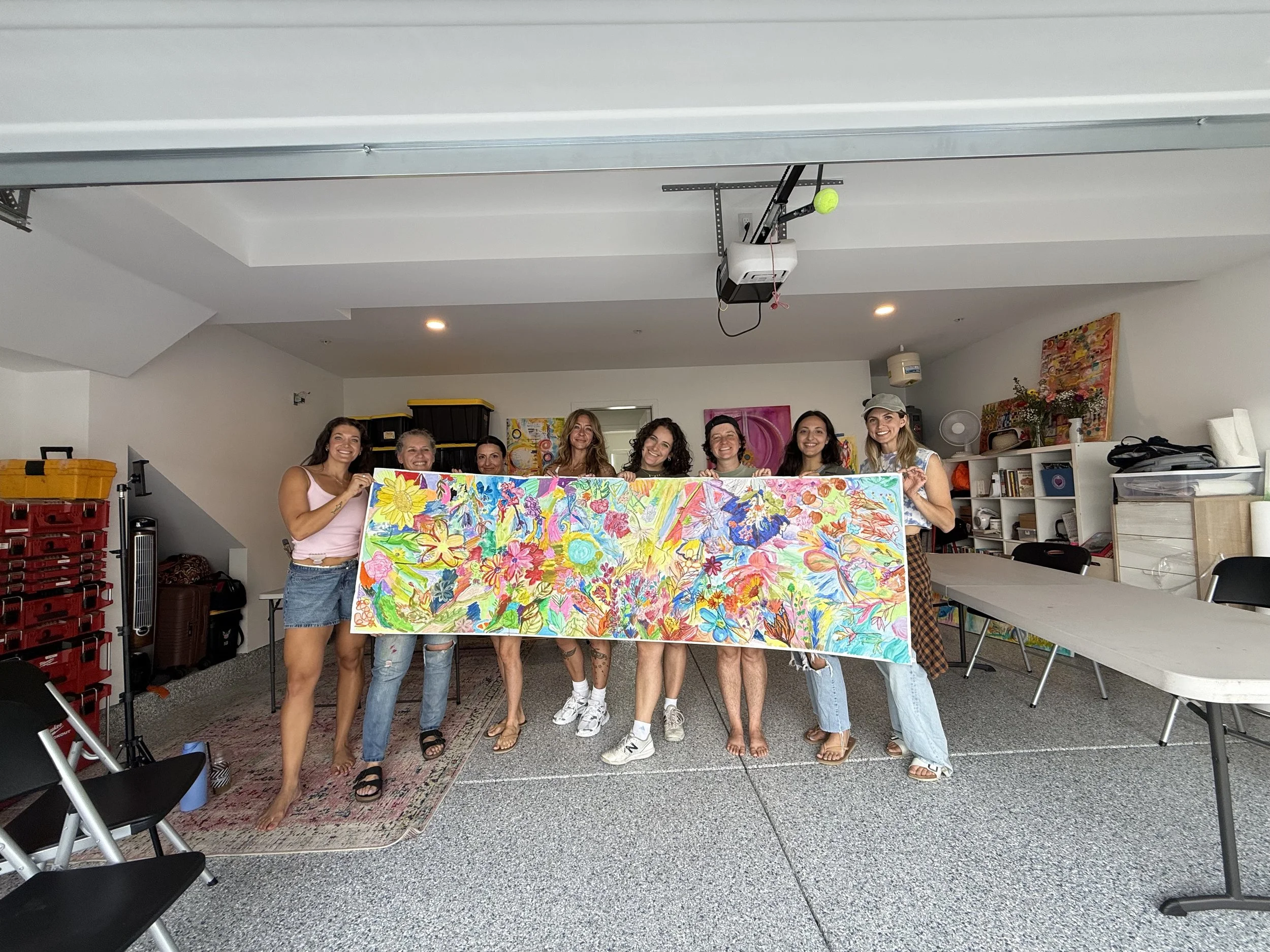 Group of eight women standing indoors, holding a large colorful floral painting, in front of a white wall with artwork and shelves, some smiling and dressed casually.