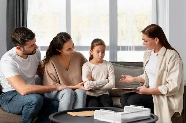 A family of three, including a man, woman, and young girl, sitting on a couch during a counseling or therapy session with a therapist, who is holding a tablet and speaking to them.
