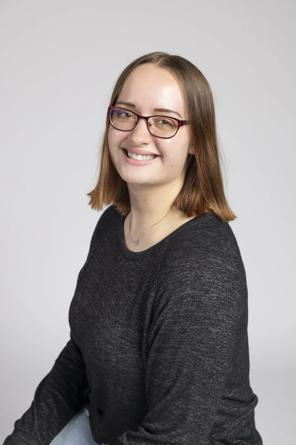 A woman smiling wearing glasses and a black sweater against a gray background.