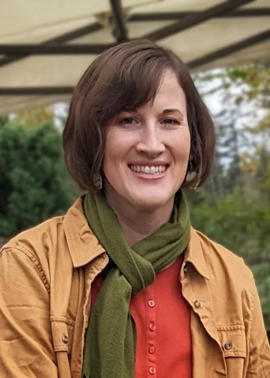 A woman smiling outdoors with trees in the background, wearing an orange shirt, a tan jacket, and a green scarf.
