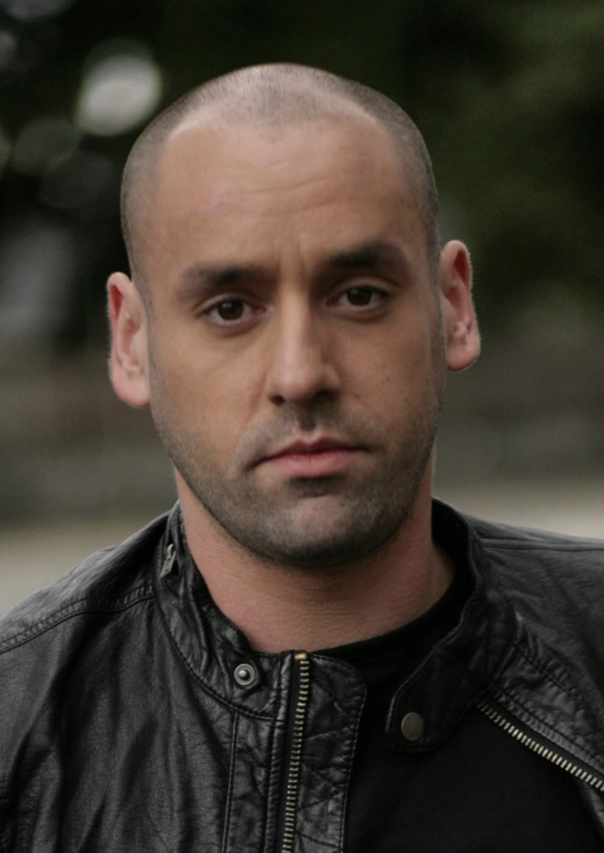 Close-up of a man with a shaved head wearing a black leather jacket, outdoors with blurred green background.