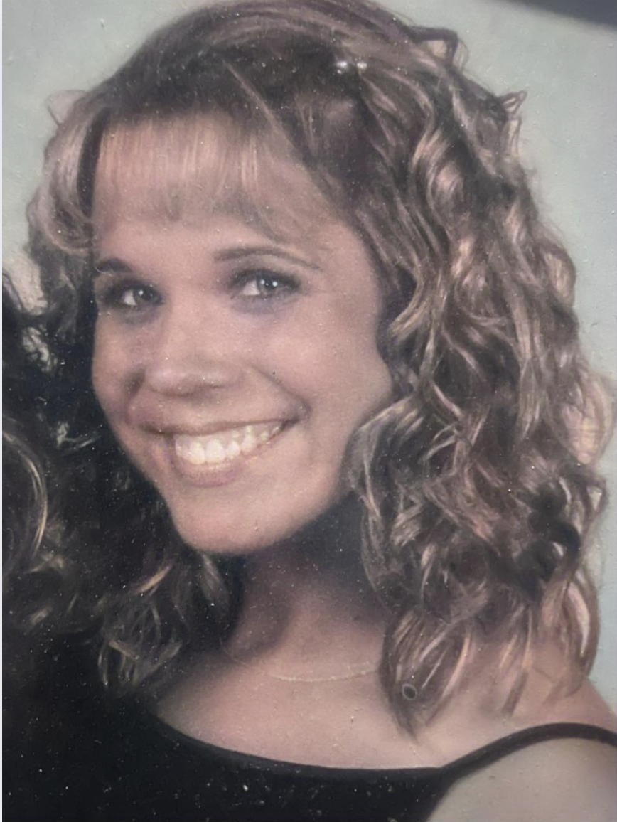 Close-up portrait of a woman with curly brown hair, smiling, wearing a black top and a thin gold necklace.