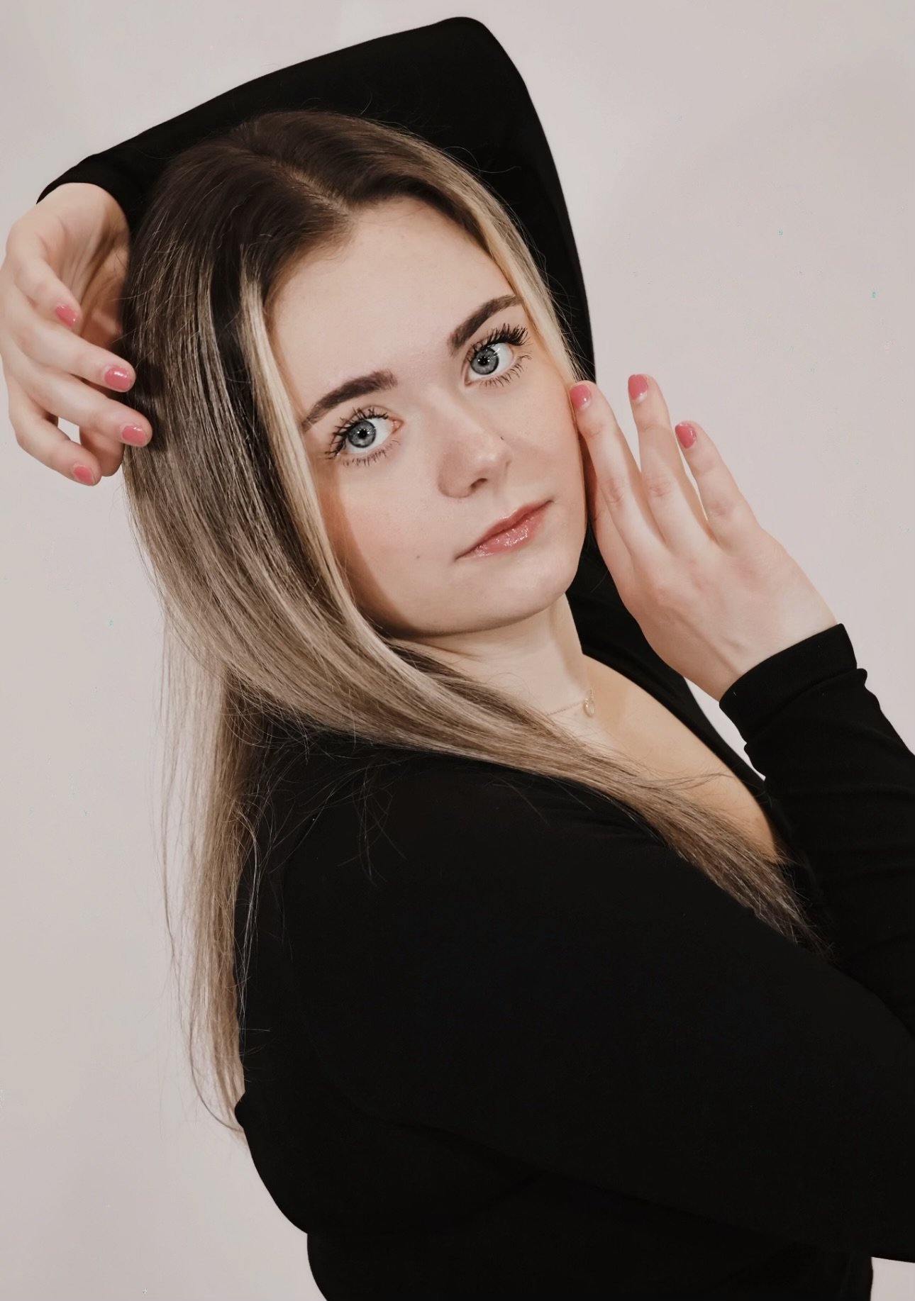 A young woman with long blonde hair, blue eyes, wearing a black top, posing with one hand on her face and the other behind her head against a plain light background.
