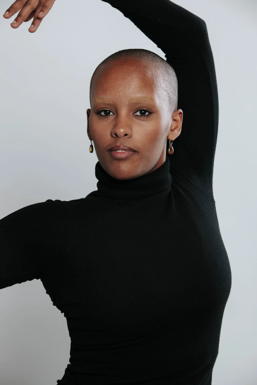 A woman with a shaved head wearing a black turtleneck and earrings, posing with one arm raised above her head against a plain background.