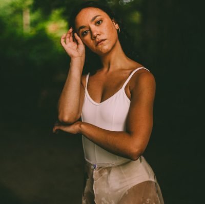 A woman standing outdoors with a forest background, wearing a white tank top and beige pants, gazing at the camera.