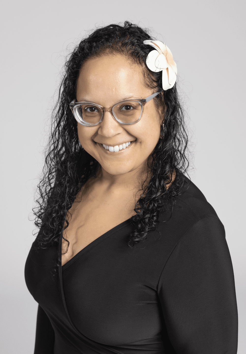 A woman with curly black hair, glasses, and a white flower in her hair, smiling, wearing a black top, black background.