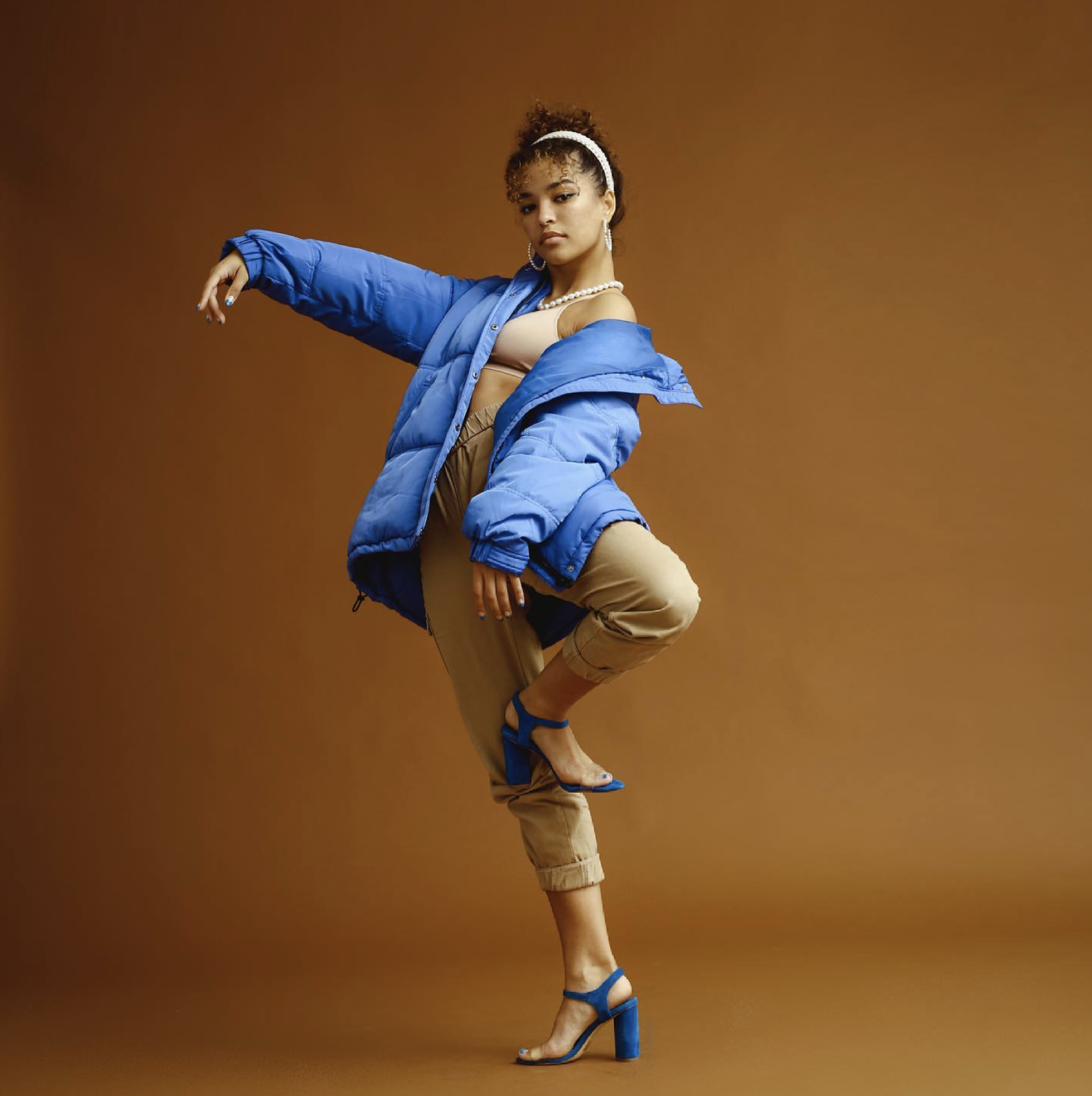A woman with curly hair wearing a blue puffer jacket, beige top, beige pants, and blue high-heeled sandals, striking a dance pose against a solid brown background.