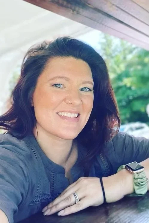 A woman with brown hair, blue eyes, and a nose ring smiling at the camera. She is sitting outdoors under a wooden roof, with greenery in the background.