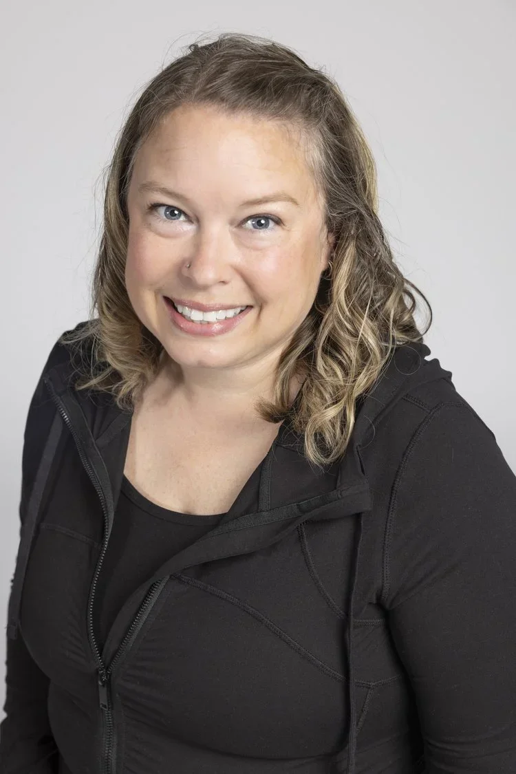 A smiling woman with curly blonde hair, wearing a black athletic jacket, posing against a plain light gray background.