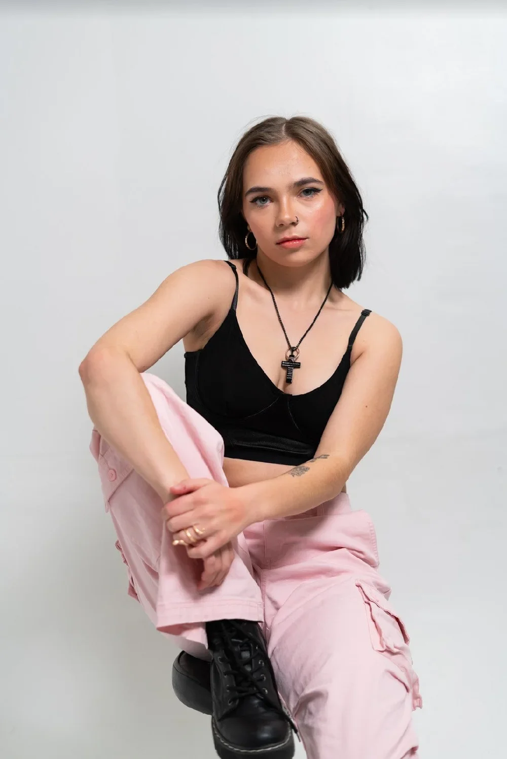 A young woman with dark brown hair and blue eyes poses sitting on one knee against a plain white background. She wears a black crop top, pink cargo pants, black boots, hoop earrings, and a necklace with a black cross pendant. She has a facial piercing and tattoos on her arm and hand.