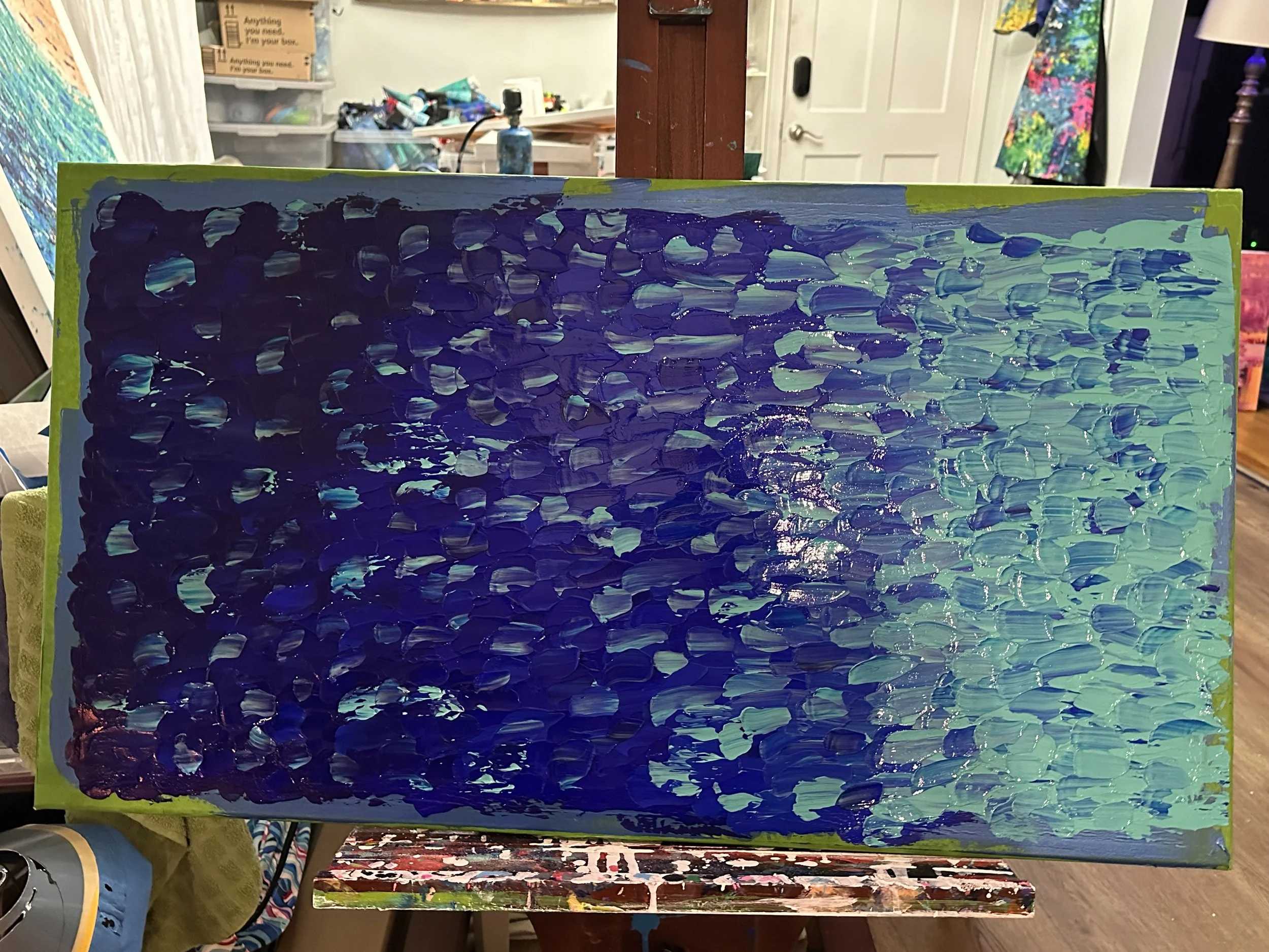 Abstract painting with brush strokes in shades of blue, purple, and hints of green on a canvas. Art supplies and clutter visible in background.