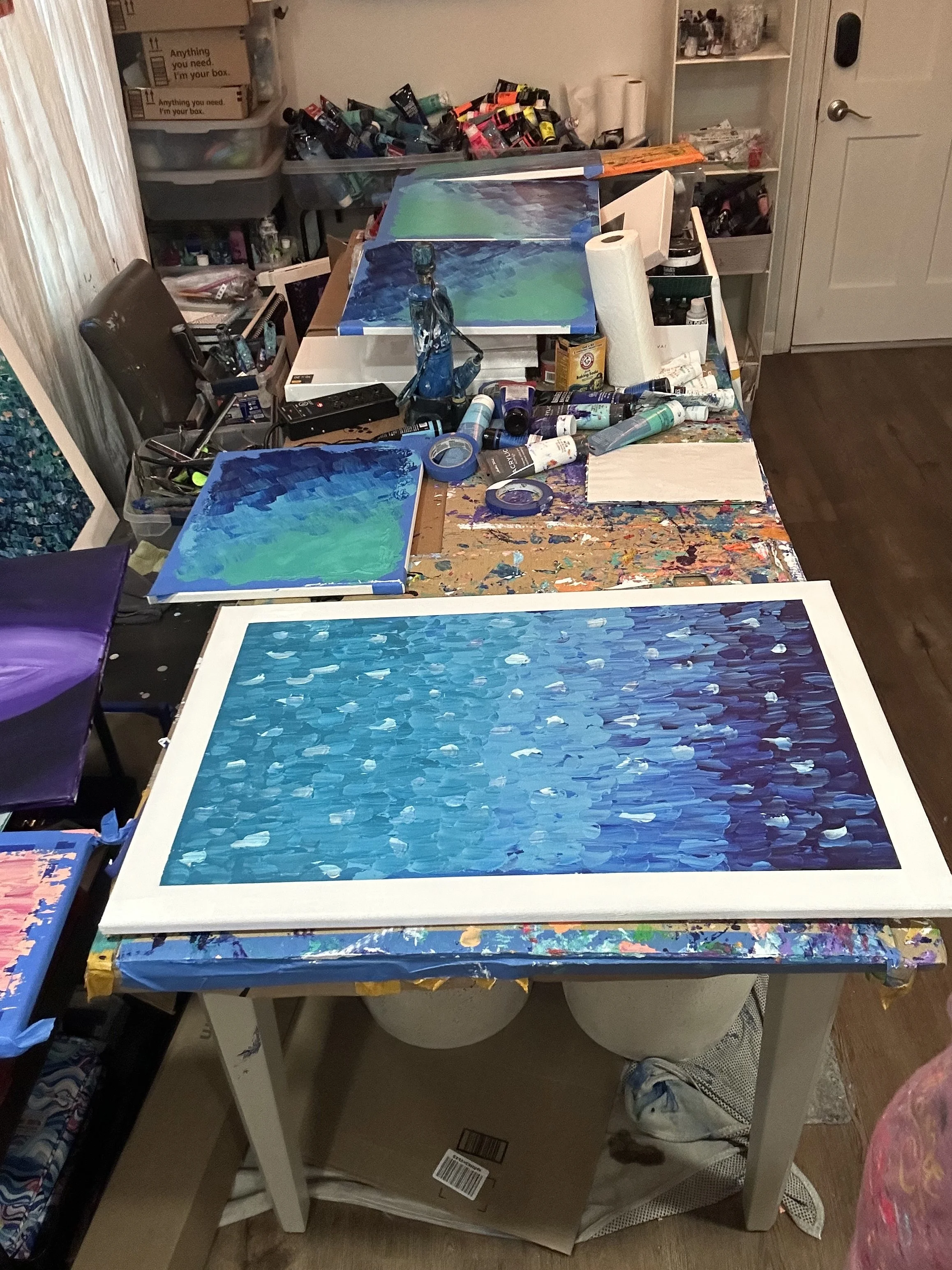 A workspace with paintings in progress on the table, surrounded by art supplies such as tubes of paint, painter's tape, and paper towels. There are organized storage containers and a chair nearby.