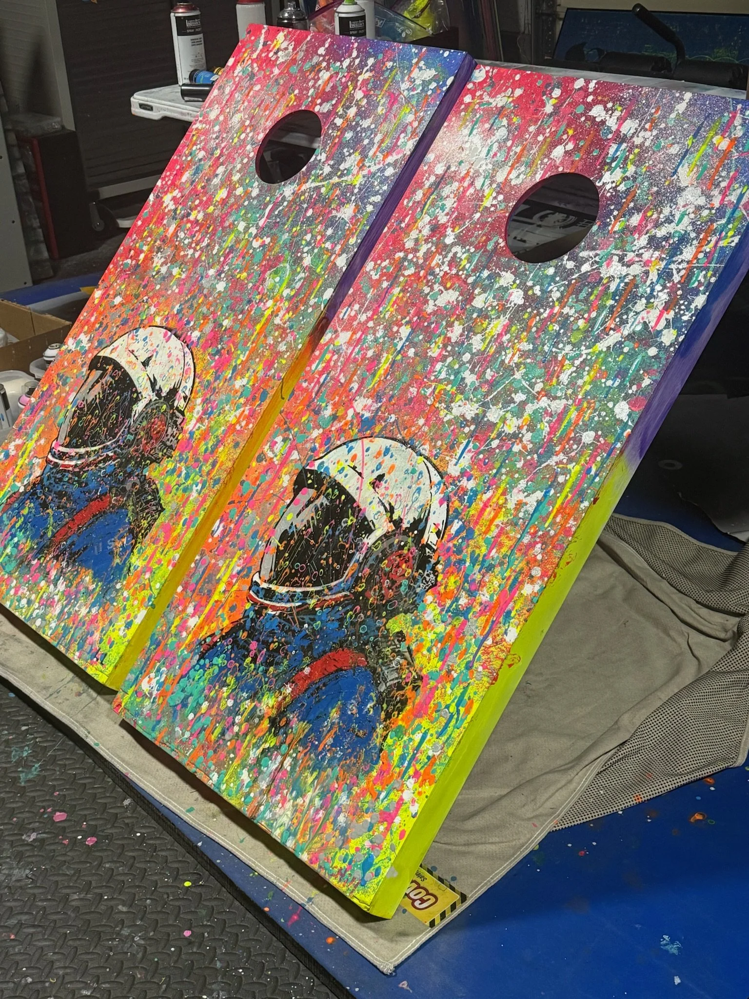 Two colorful, splatter-painted cornhole boards with images of astronaut helmets, placed on a worktable in an art studio or workshop.