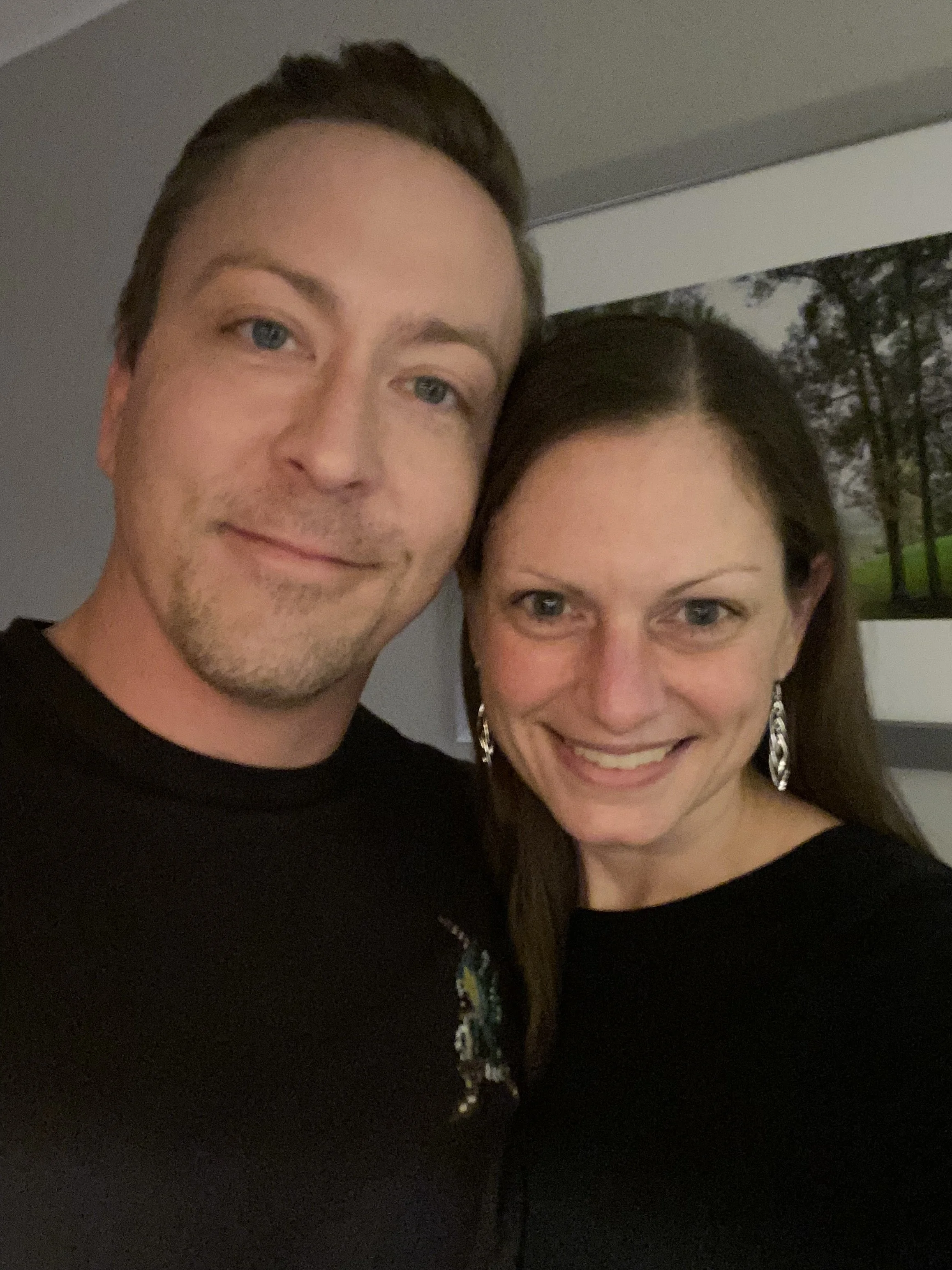 A close-up of a man and woman smiling for a selfie indoors, with a painting of trees in the background.