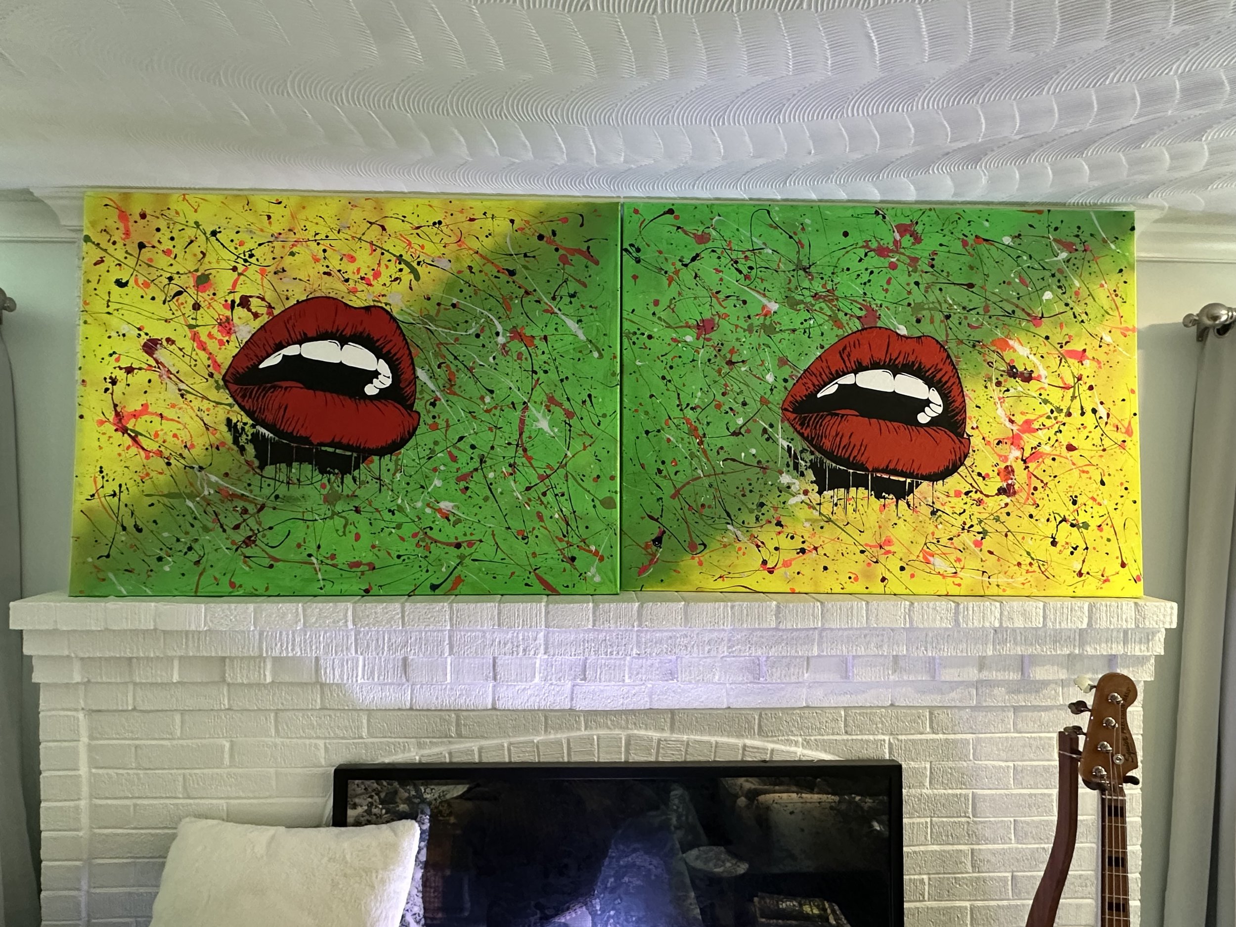 A colorful abstract painting of two open mouths with red lips and white teeth, set against a yellow and green splatter background, hung above a white painted brick fireplace.