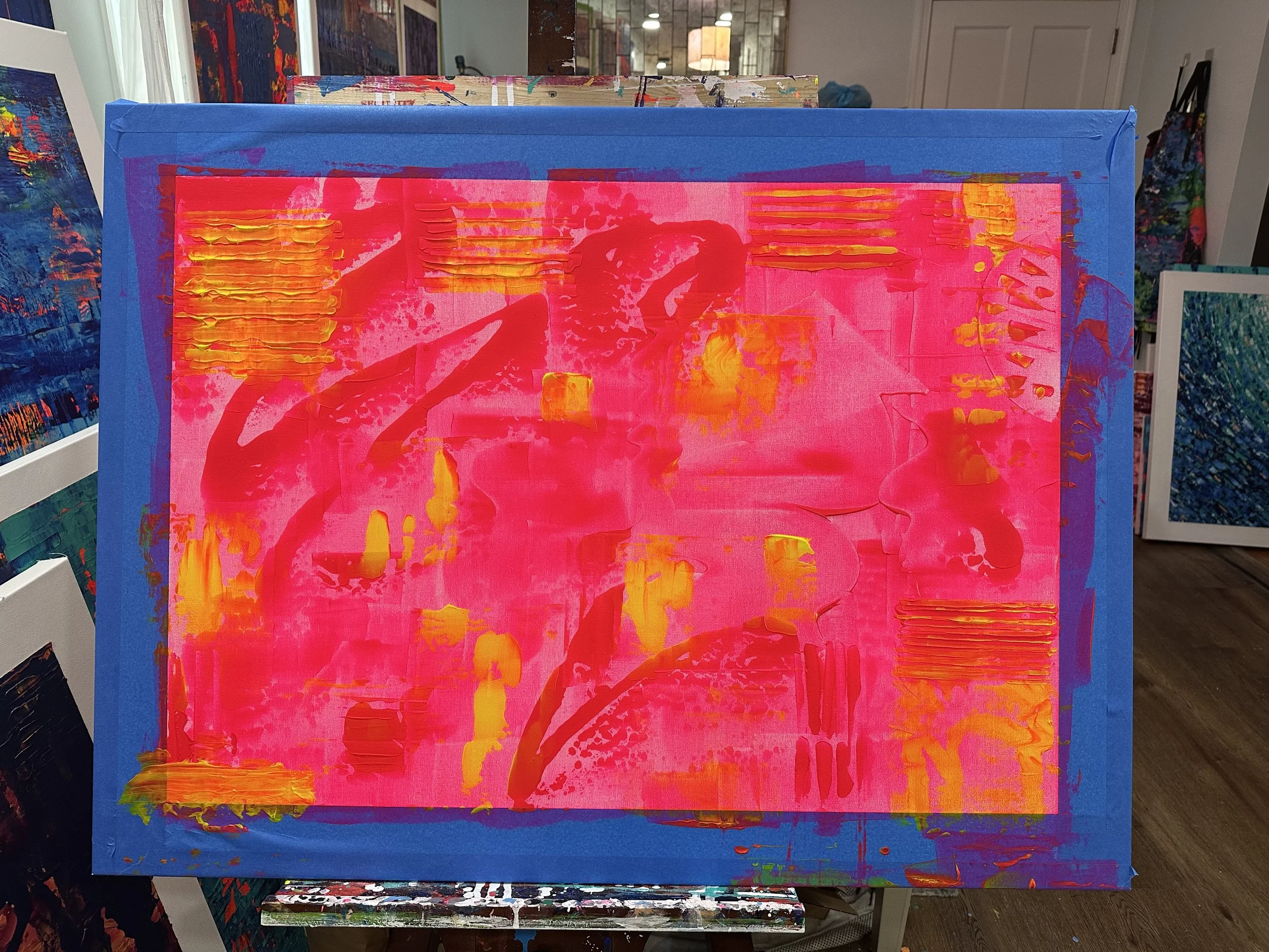 Abstract acrylic painting with a bright hot pink background, orange and yellow highlights, and blue borders, displayed in an art gallery with other colorful artworks in the background.