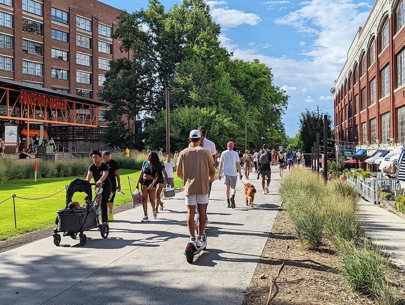 People walking on the belt line in Atlanta on a sunny day, some with strollers and backpacks, alongside tall buildings and greenery.