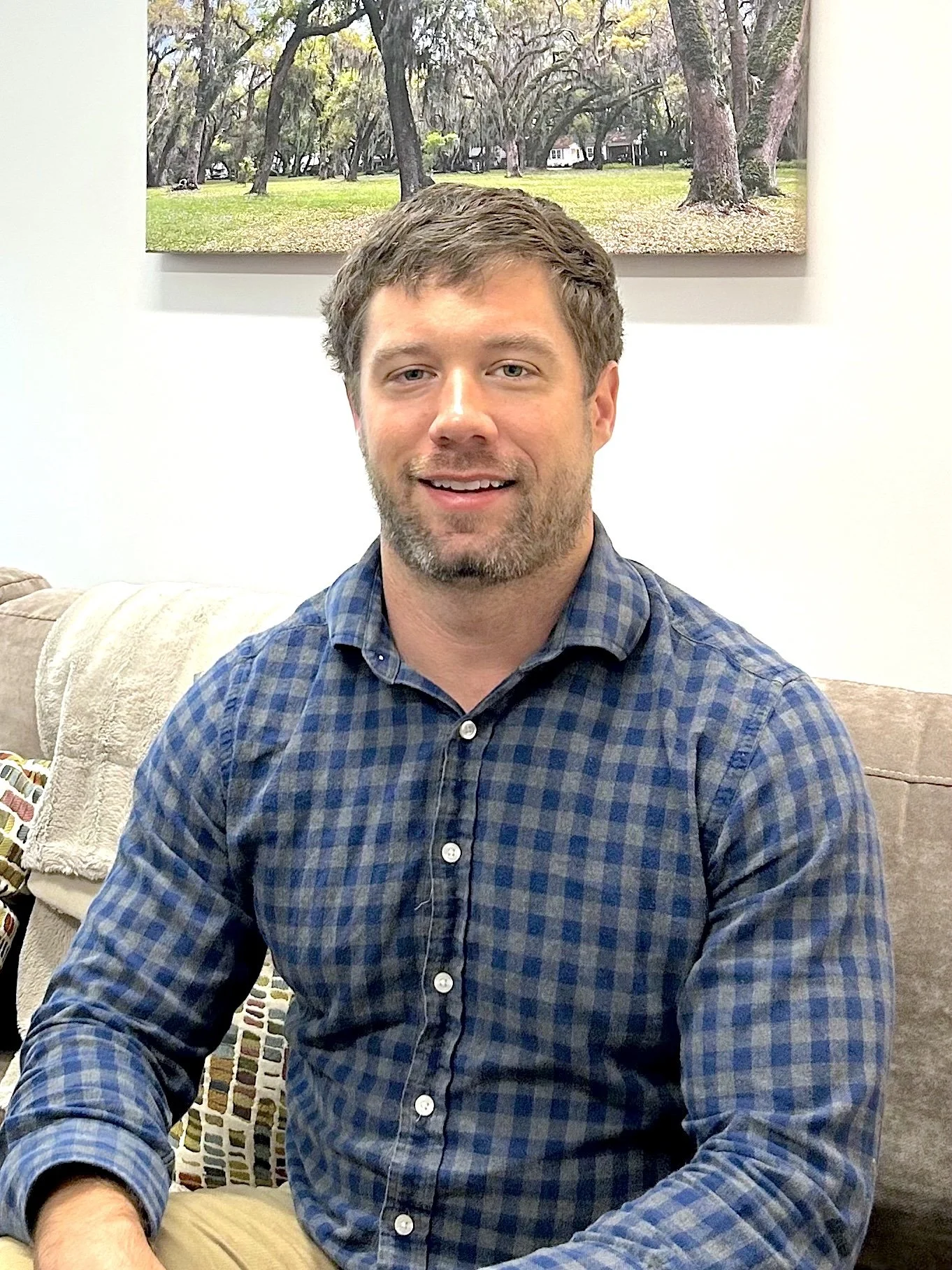 A man, Palmer McCluskey, with short brown hair and a beard, wearing a blue and black checkered shirt, sitting on a beige couch in a room with white walls and a landscape painting of trees hanging behind him. He is smiling and looking at the camera.