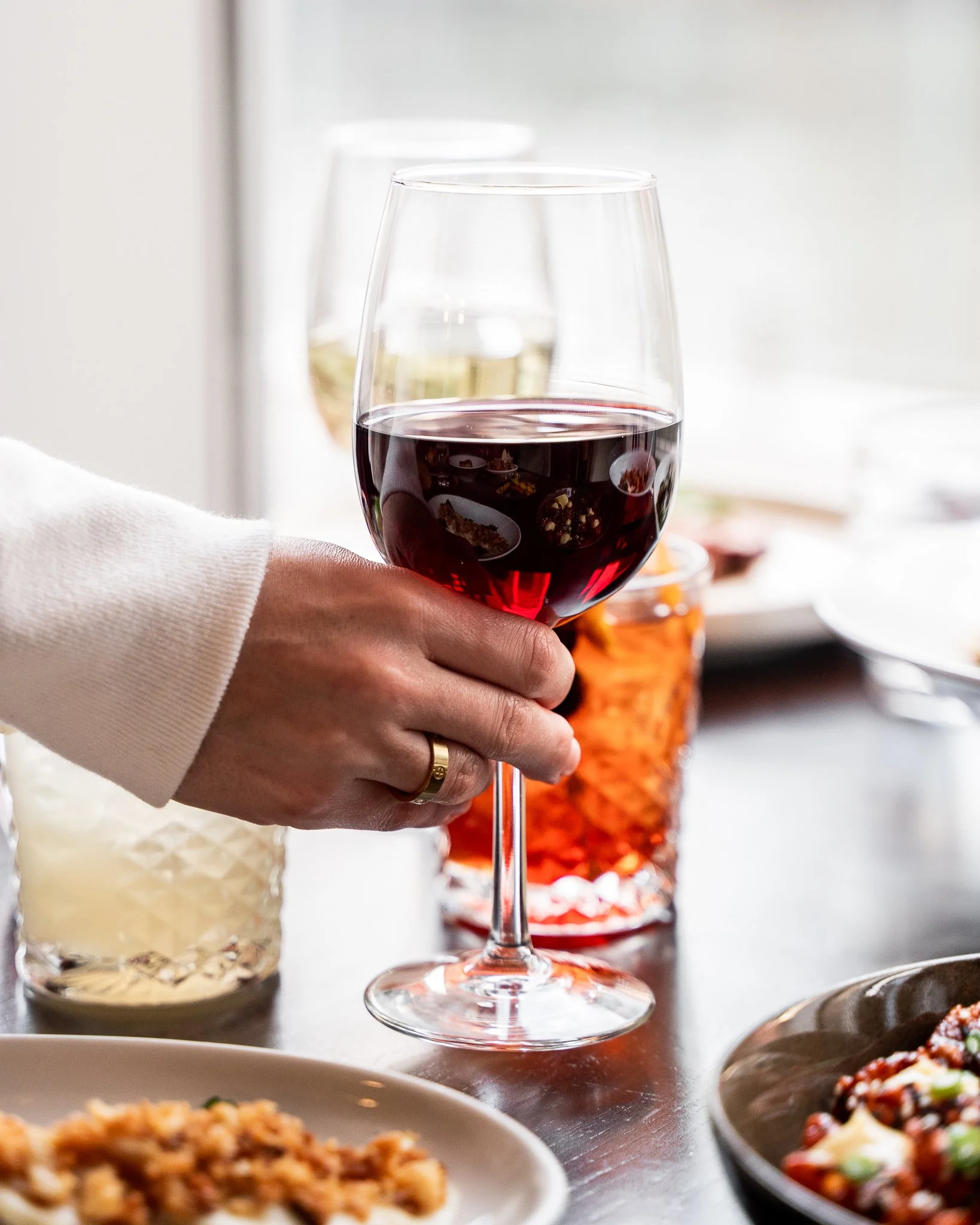 Hand holding a glass of red wine near a table with plates of food, including pasta and a salad, in a well-lit dining area.