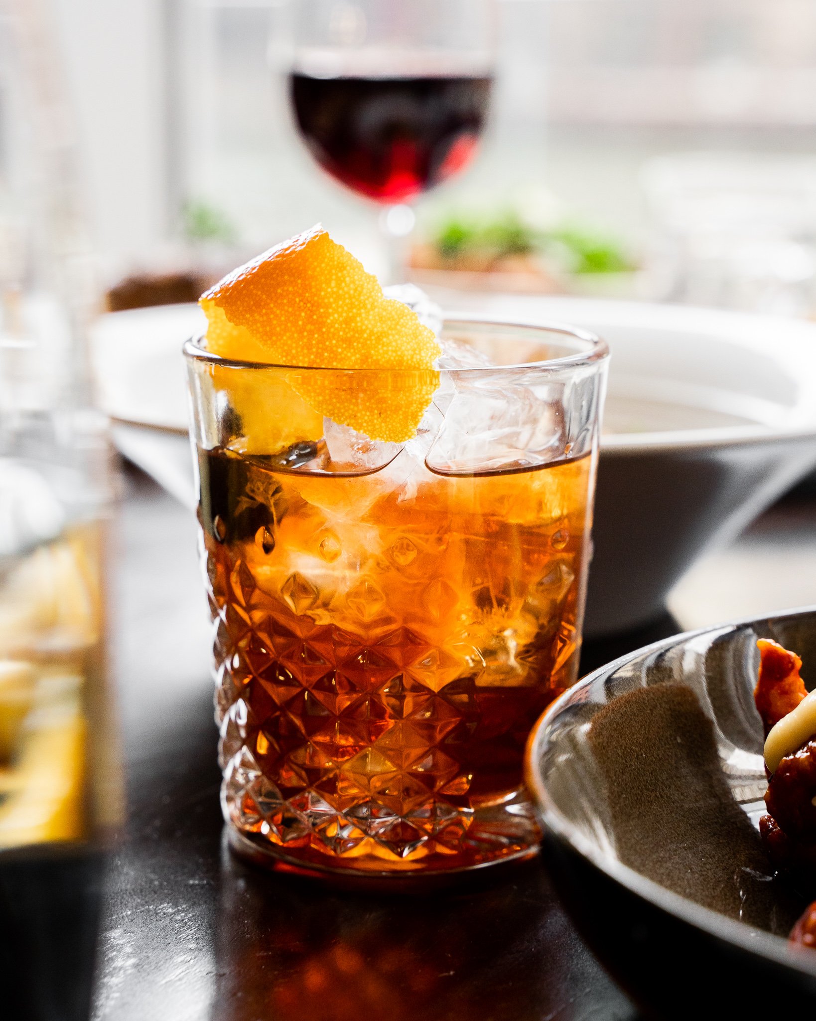 A glass of amber-colored beverage with ice and a slice of orange on top, with a glass of red wine in the background and a blurry plate of food to the side.