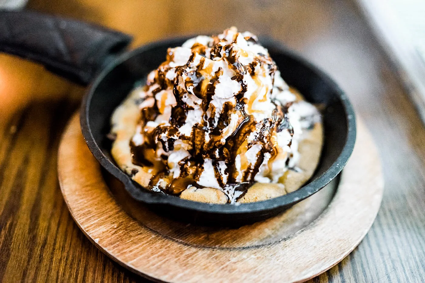 A dessert in a small black cast iron skillet topped with whipped cream, caramel drizzle, and chocolate syrup, set on a round wooden serving board.