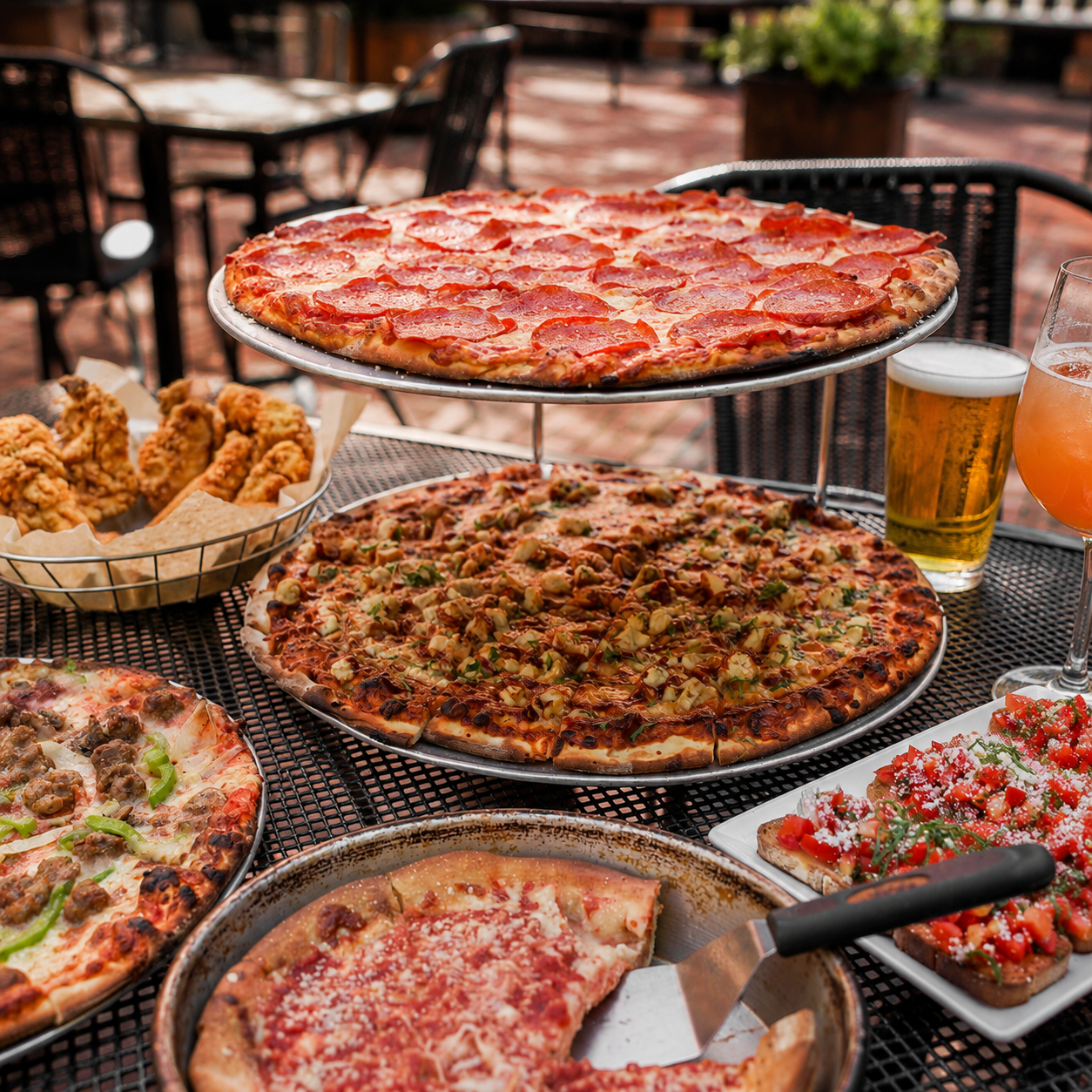 Assorted pizzas including pepperoni, vegetable, and specialty toppings, along with fried chicken, salads, and beverages at an outdoor dining table.
