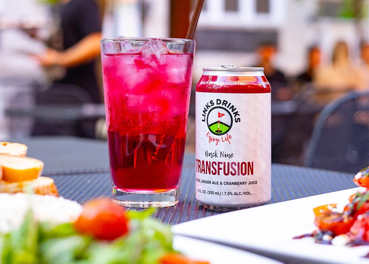 A glass of pinkish-red beverage with ice and a straw next to a can labeled 'Link's Drinks Transfusion' with ginger ale and cranberry juice, on a table with blurred background of people.